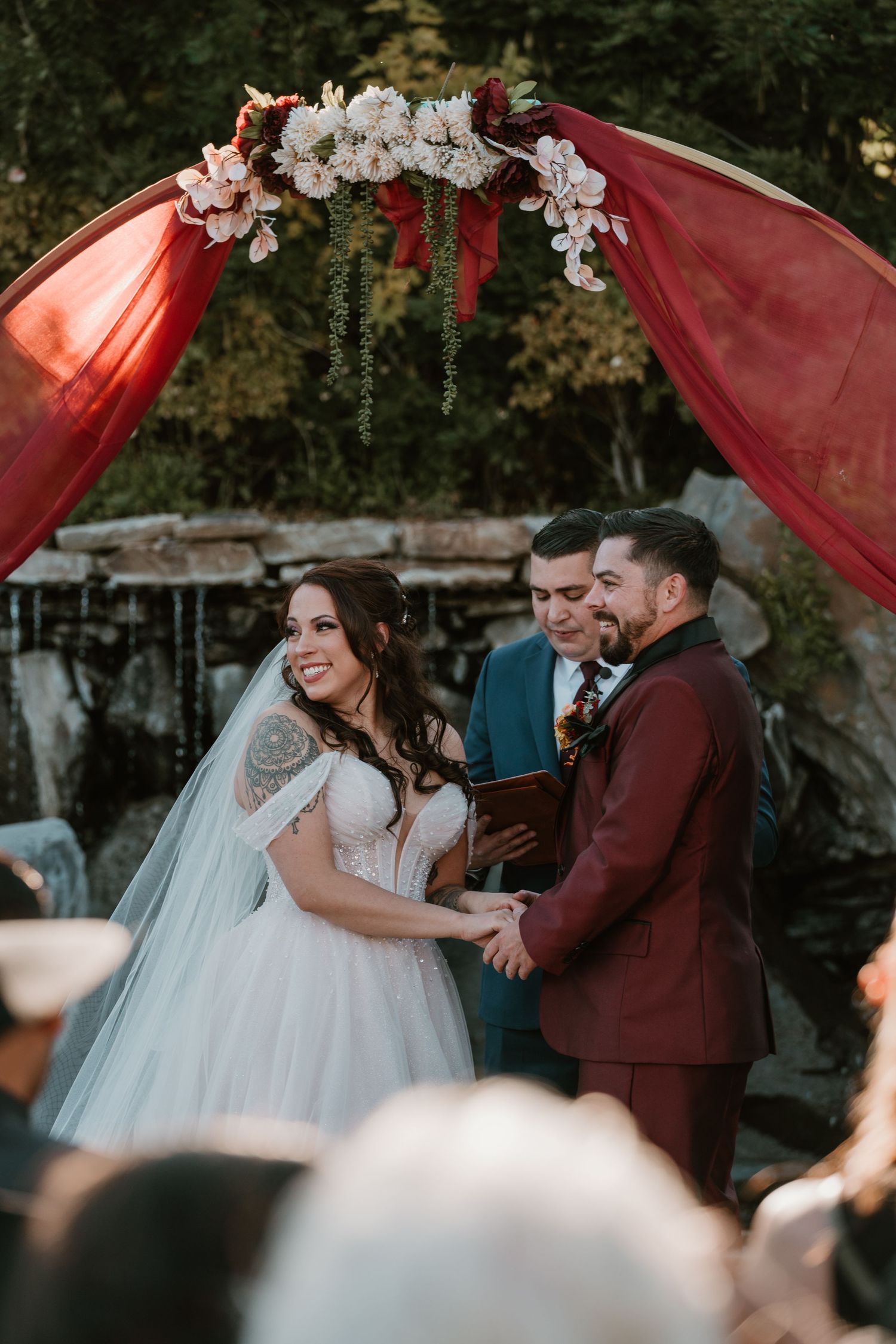 A couple shares romantic moments during an outdoor wedding ceremony under flowing burgundy drapery and white flowers.