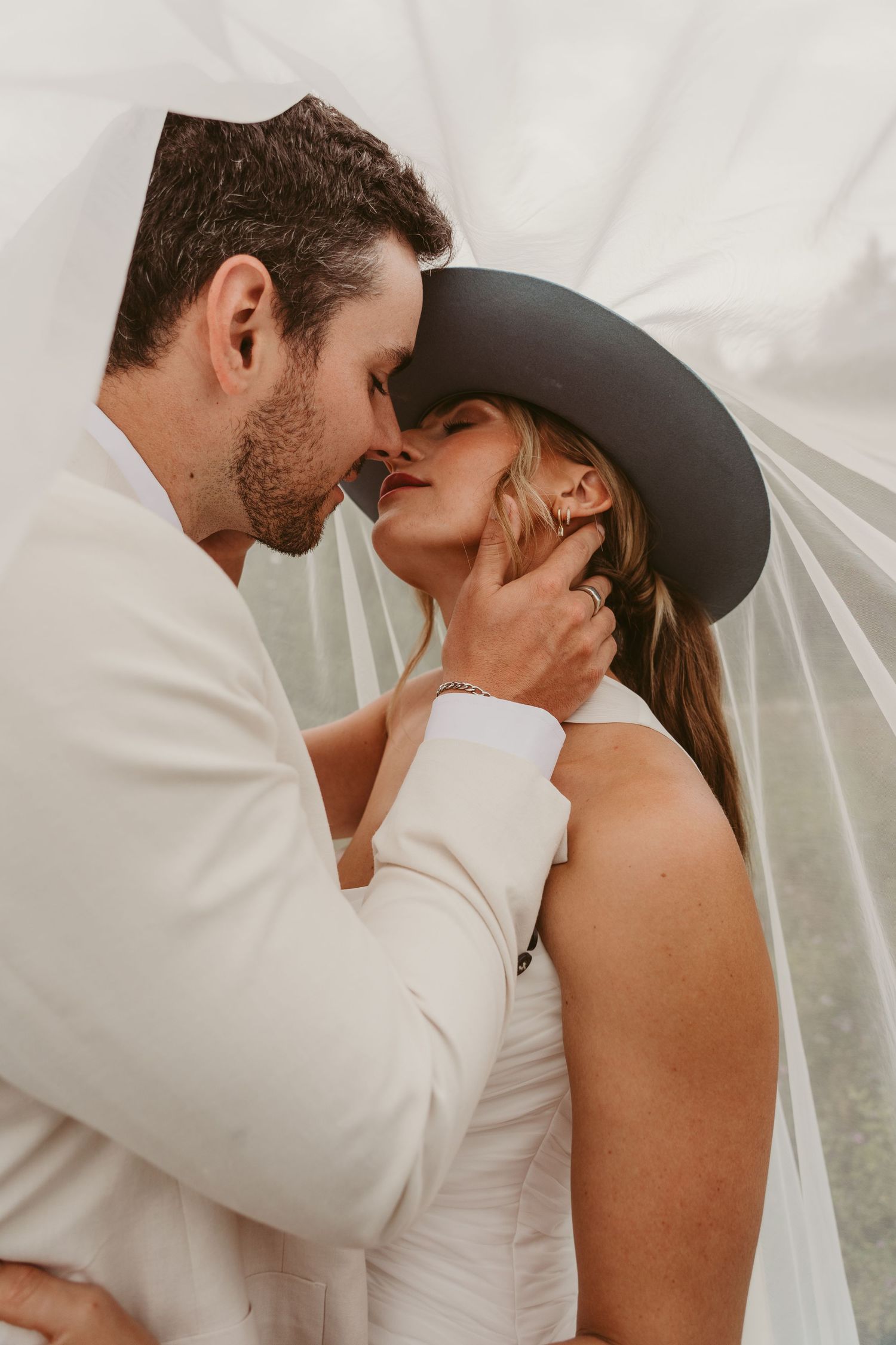 A romantic wedding couple embraces tenderly under a flowing white veil during an intimate photo session.