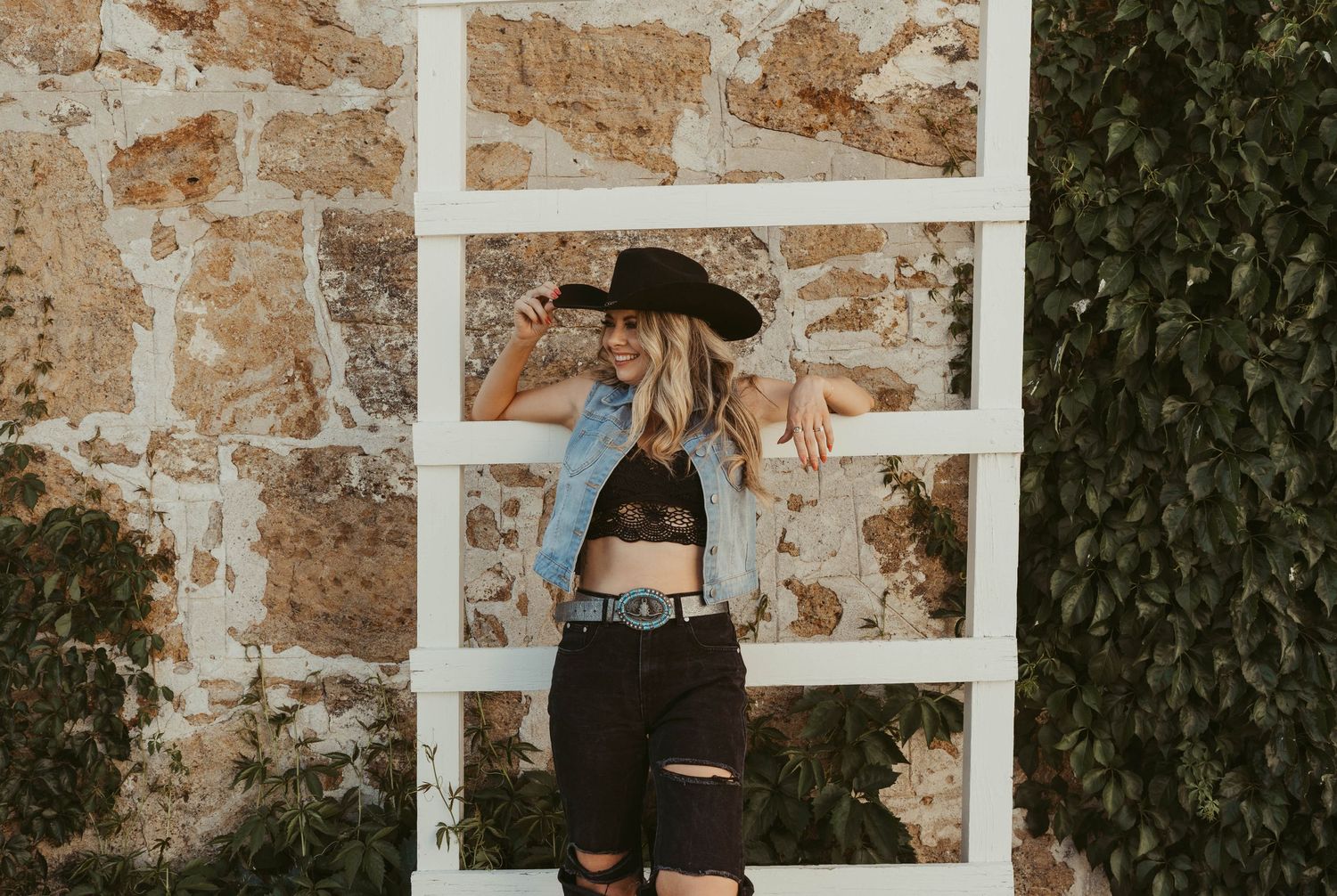 A person in distressed black jeans and a crop top poses against a stone wall with a white ladder and green vines.