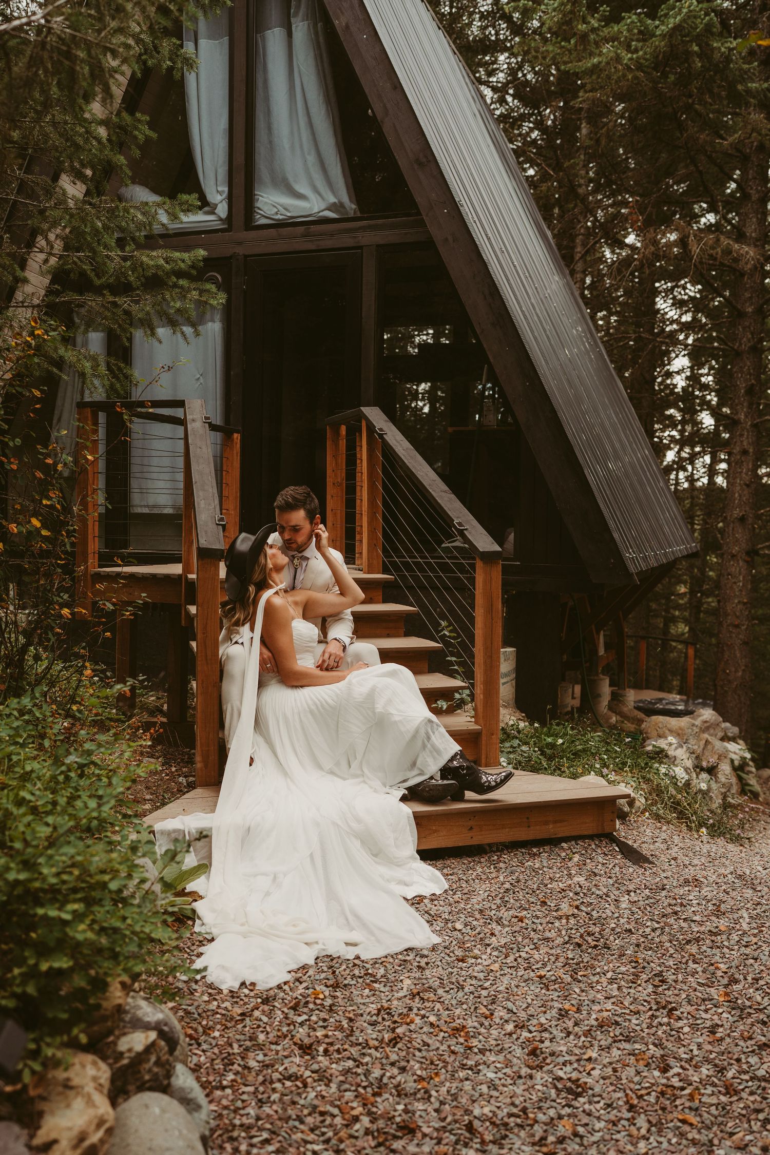 Couple in wedding attire embraces on rustic gravel path by wooden cabin in forest setting.