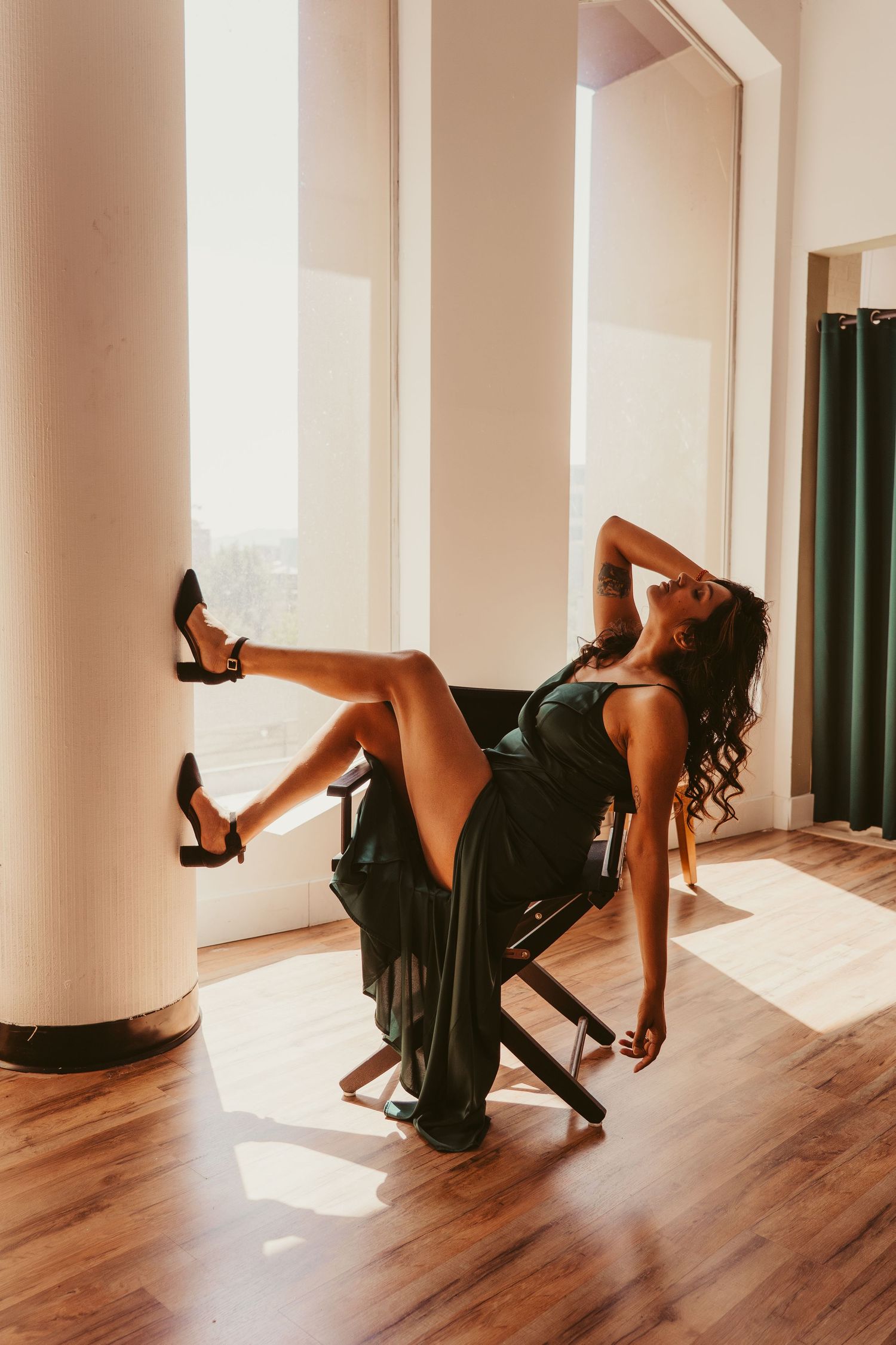 A person in a black dress and heels poses dramatically on a chair in a sunlit room with wooden floors.