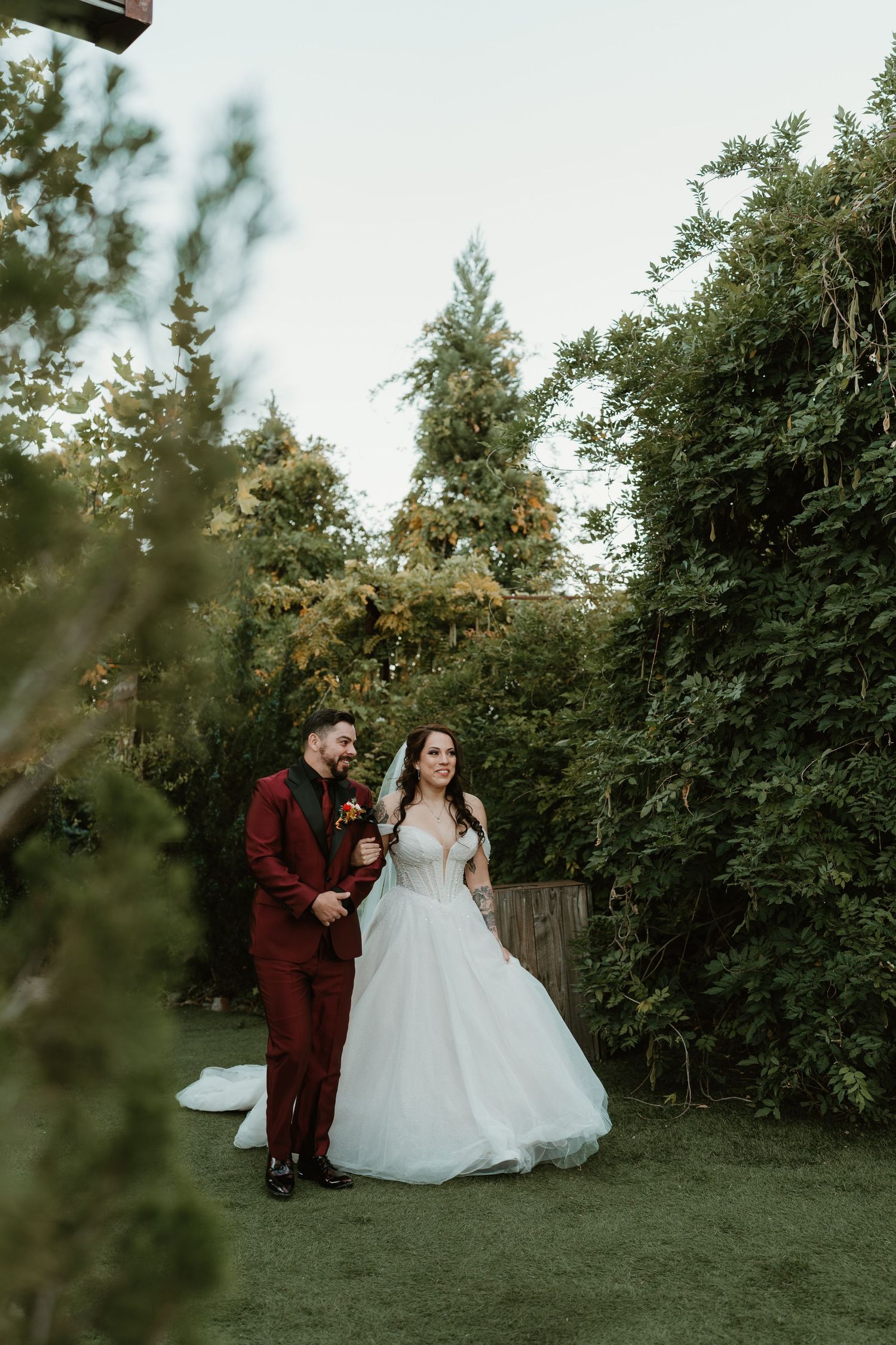 A couple in burgundy suit and white wedding dress pose together in a garden setting at sunset.