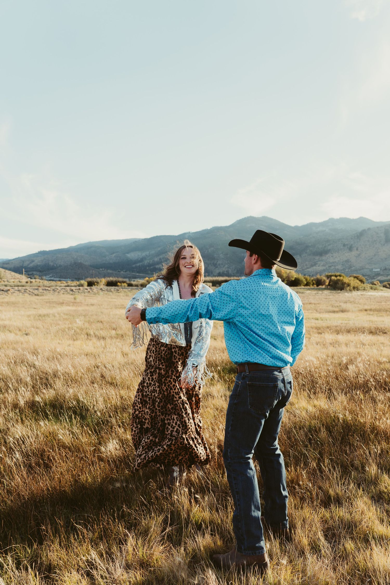 Couple dancing in golden field with mountains, woman in leopard skirt and fringe top, man in turquoise shirt and cowboy hat.