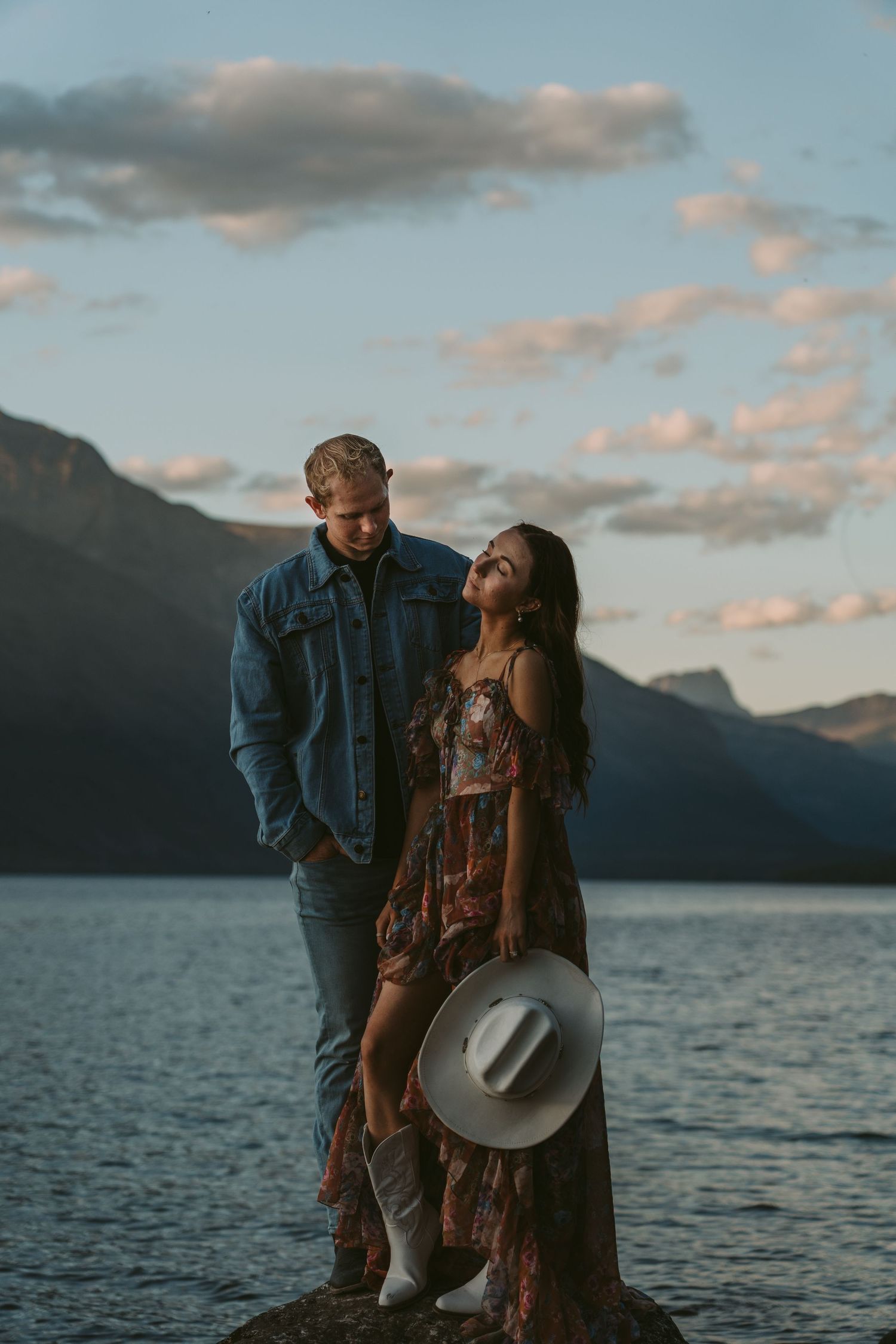 A couple shares a romantic moment by a scenic mountain lake at sunset with dramatic clouds in the background.
