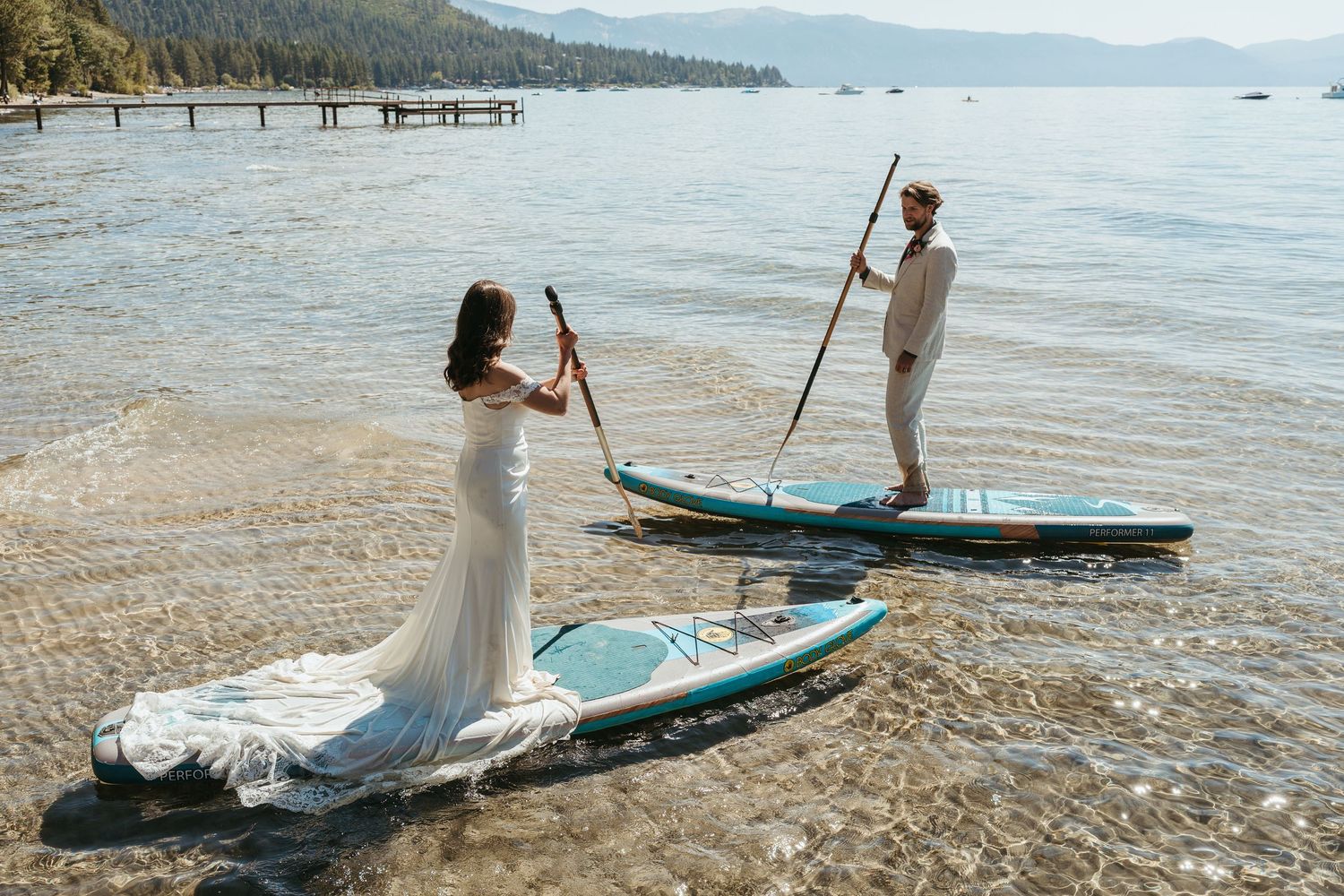 Couple in wedding attire paddling stand-up paddleboards on calm lake waters with mountains in background.