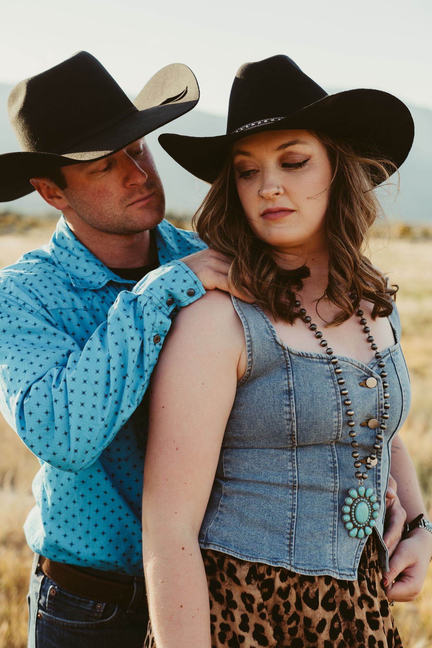 A couple in black cowboy hats and western attire pose in a golden field, man in blue shirt, woman in denim vest.