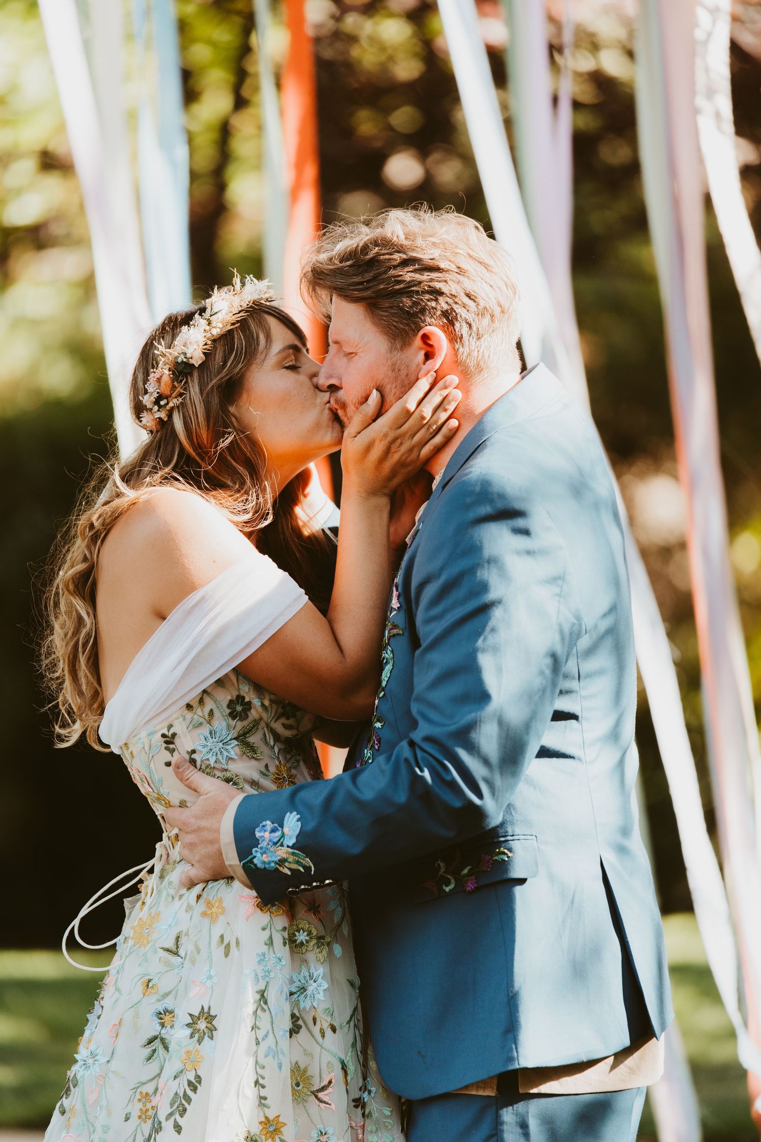 Couple sharing first kiss at outdoor bohemian wedding, bride in floral dress with flower crown, groom in blue suit.