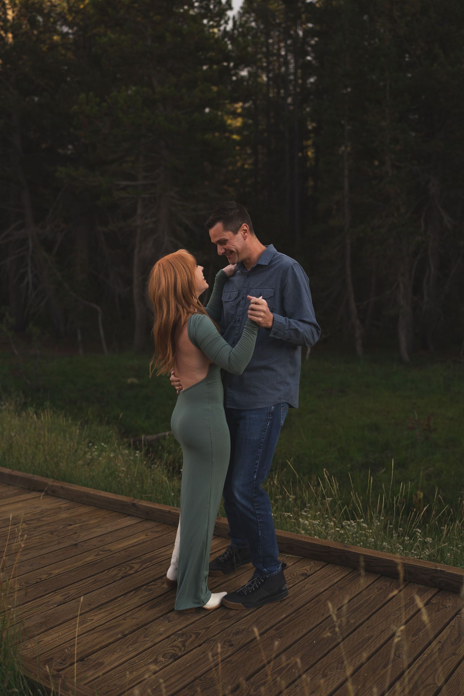 Couple dancing on wooden boardwalk at dusk near Lake Tahoe, woman in green dress, man in blue shirt, forest background.