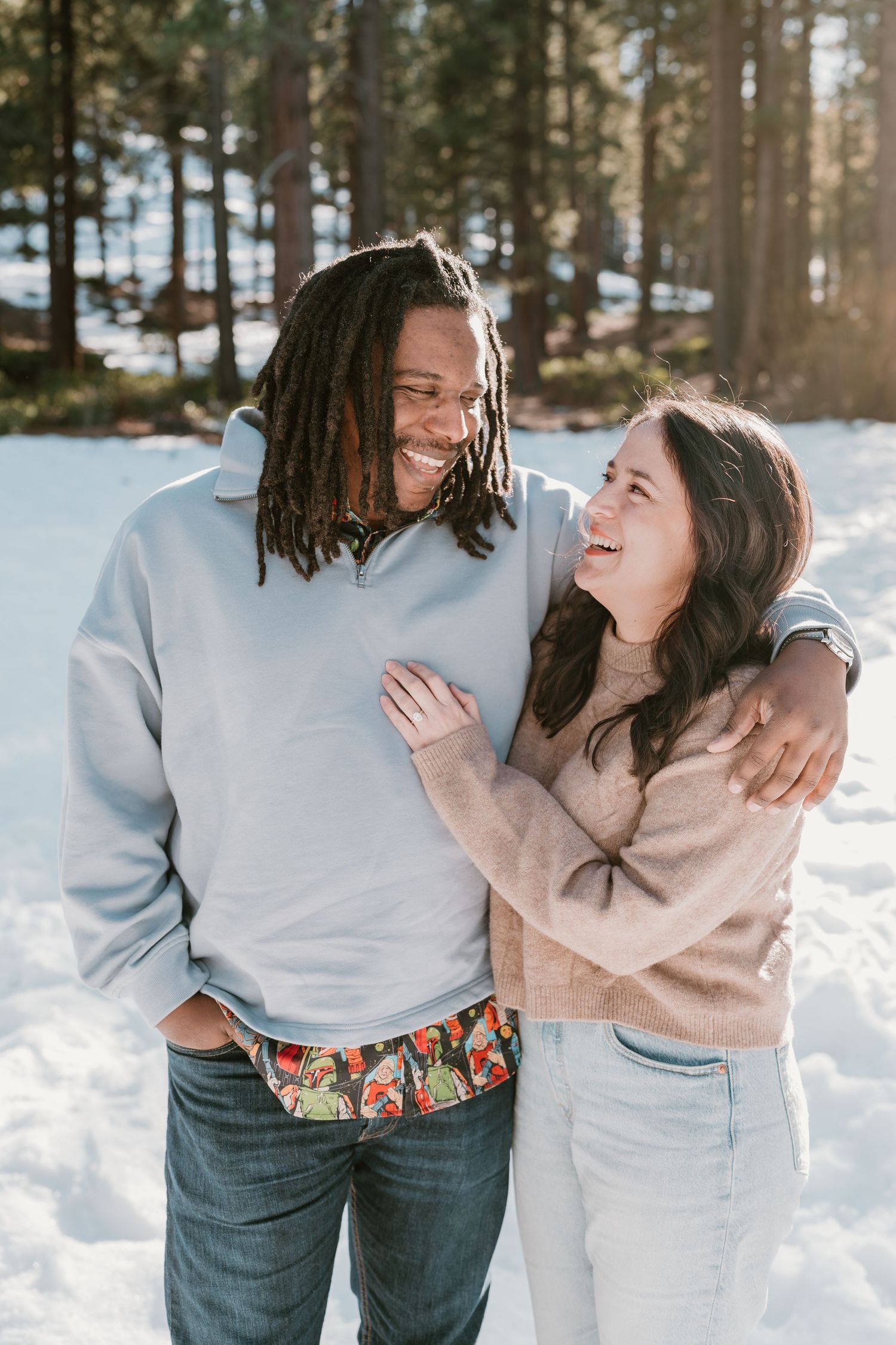 A smiling couple embraces in a snowy forest, wearing casual winter sweaters, sharing a joyful moment outdoors.