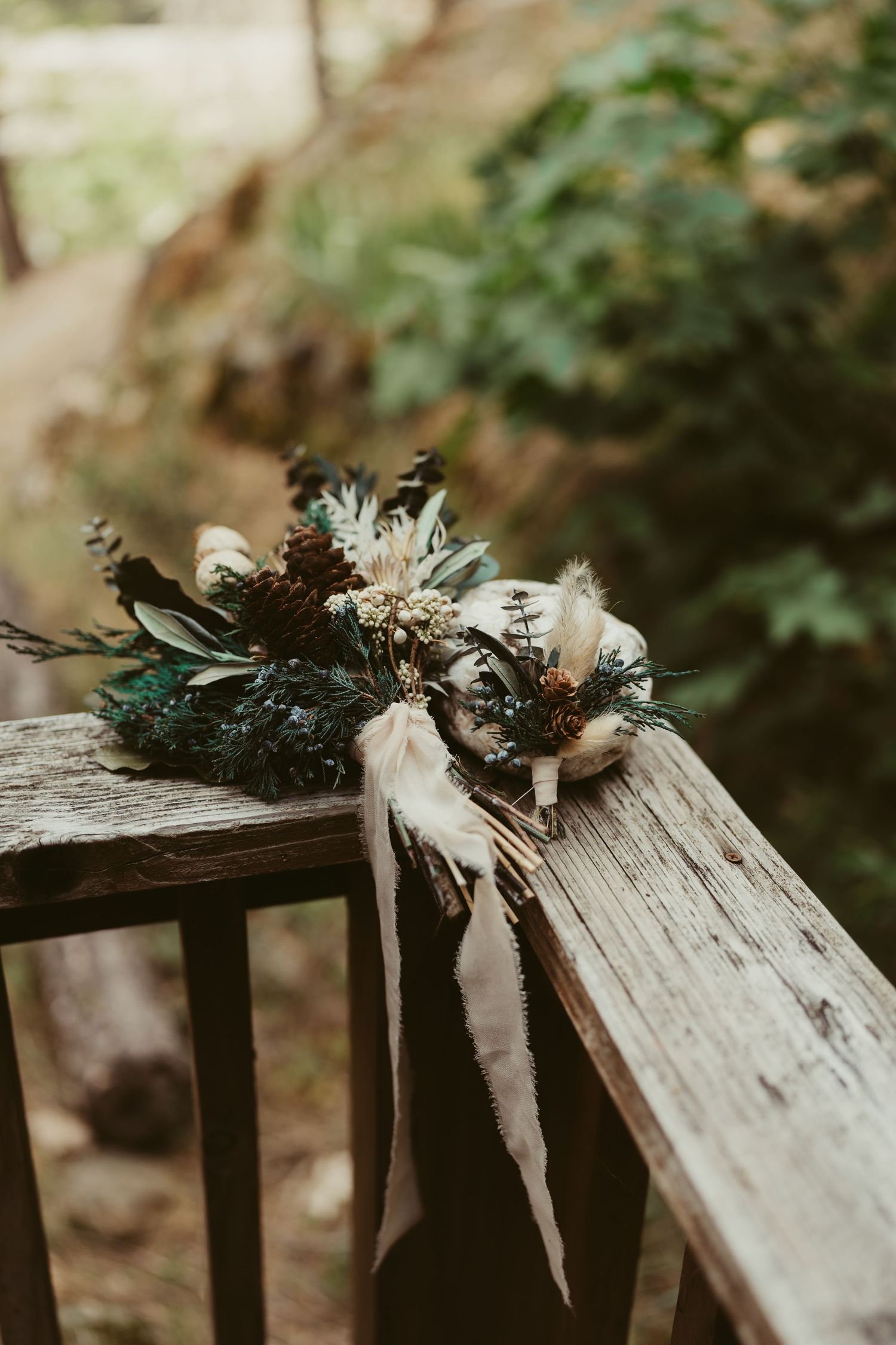 A rustic floral arrangement with dried flowers and ribbons rests on a wooden railing in a moody outdoor setting.