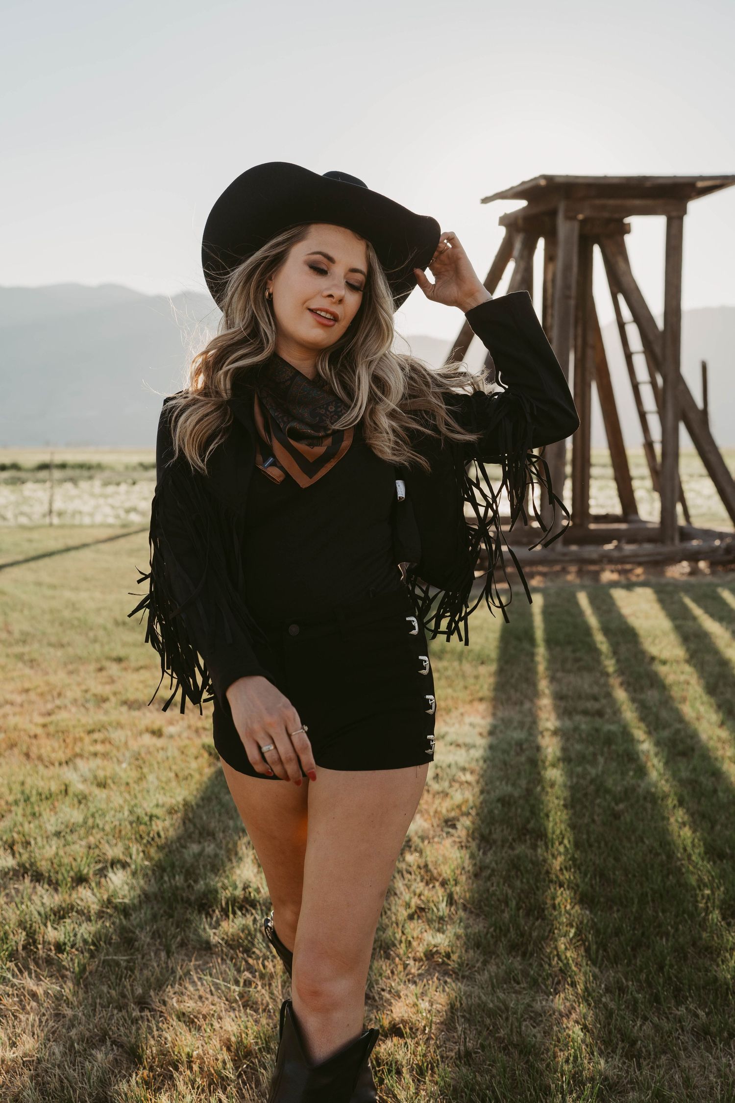 Western style outfit with black fringe jacket, shorts and hat posed in sunlit field with wooden structure backdrop.