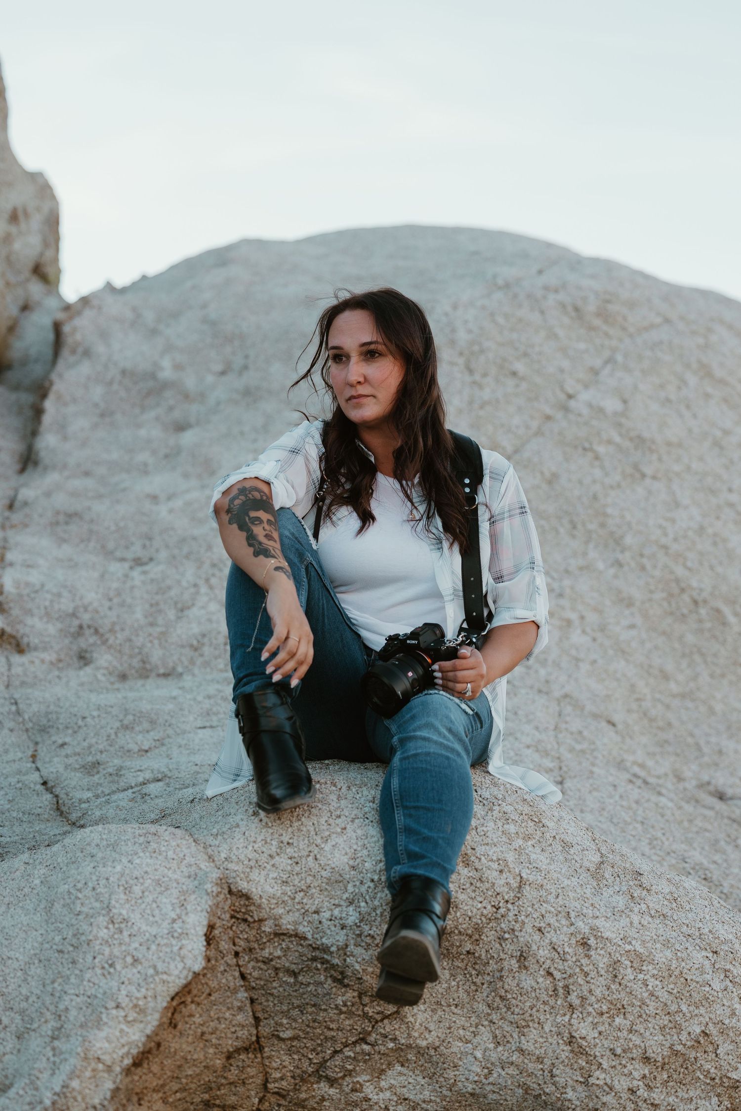 A tattooed woman sits on a boulder holding a camera, wearing jeans and a white shirt in an outdoor rocky setting.