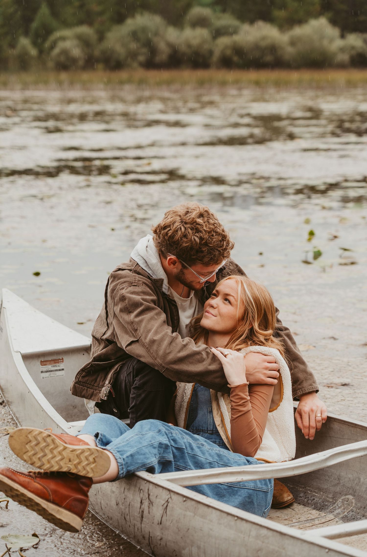 A romantic couple lounges together in a canoe by the water on an overcast day.