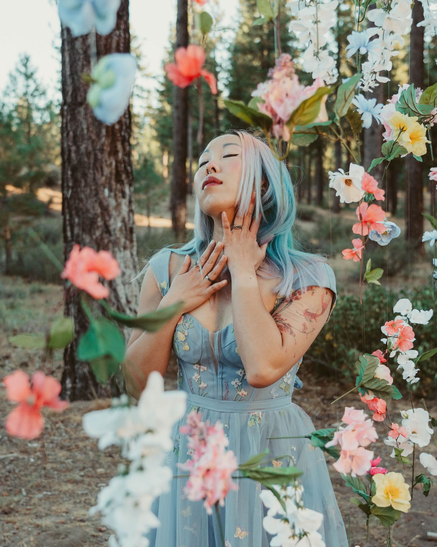 Ethereal scene in a forest with floating pink and white flowers while someone poses in a light blue dress.