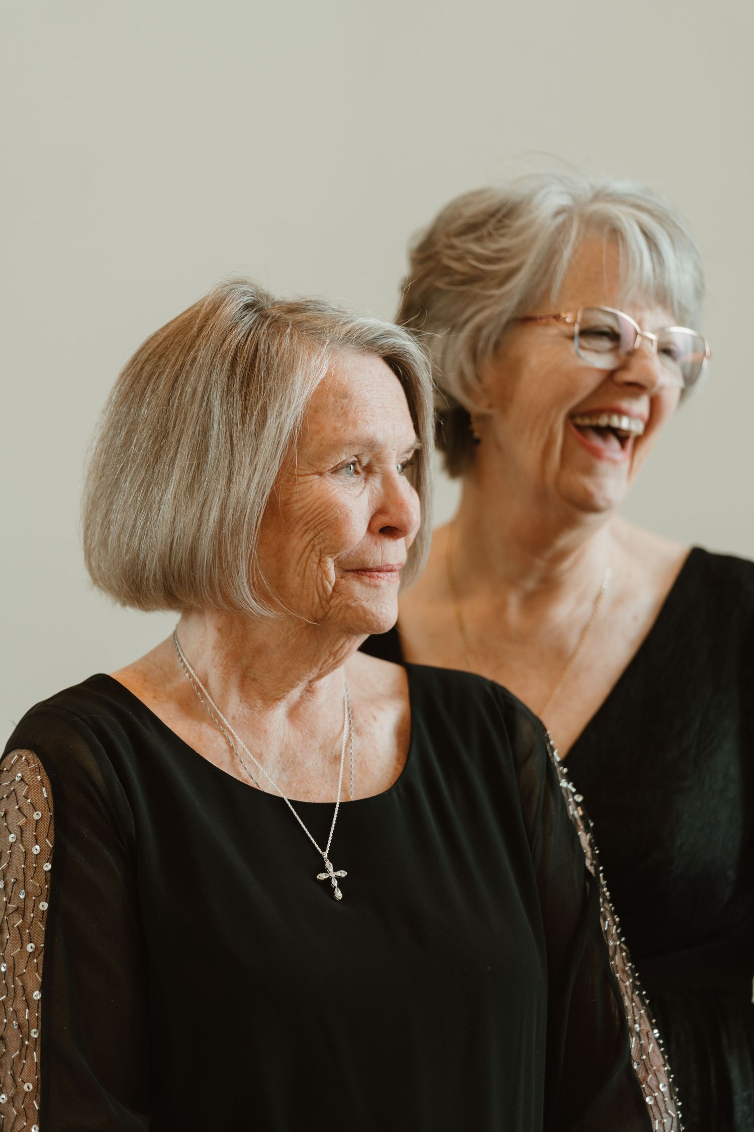 Two elegant senior women in black dresses, one wearing a cross necklace, the other laughing and wearing glasses.