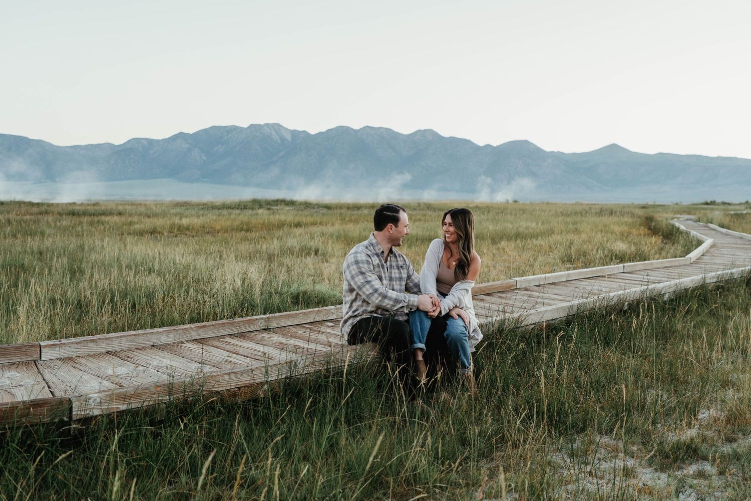 A couple sits together in tall grass beside a winding mountain road at sunset in a serene landscape.