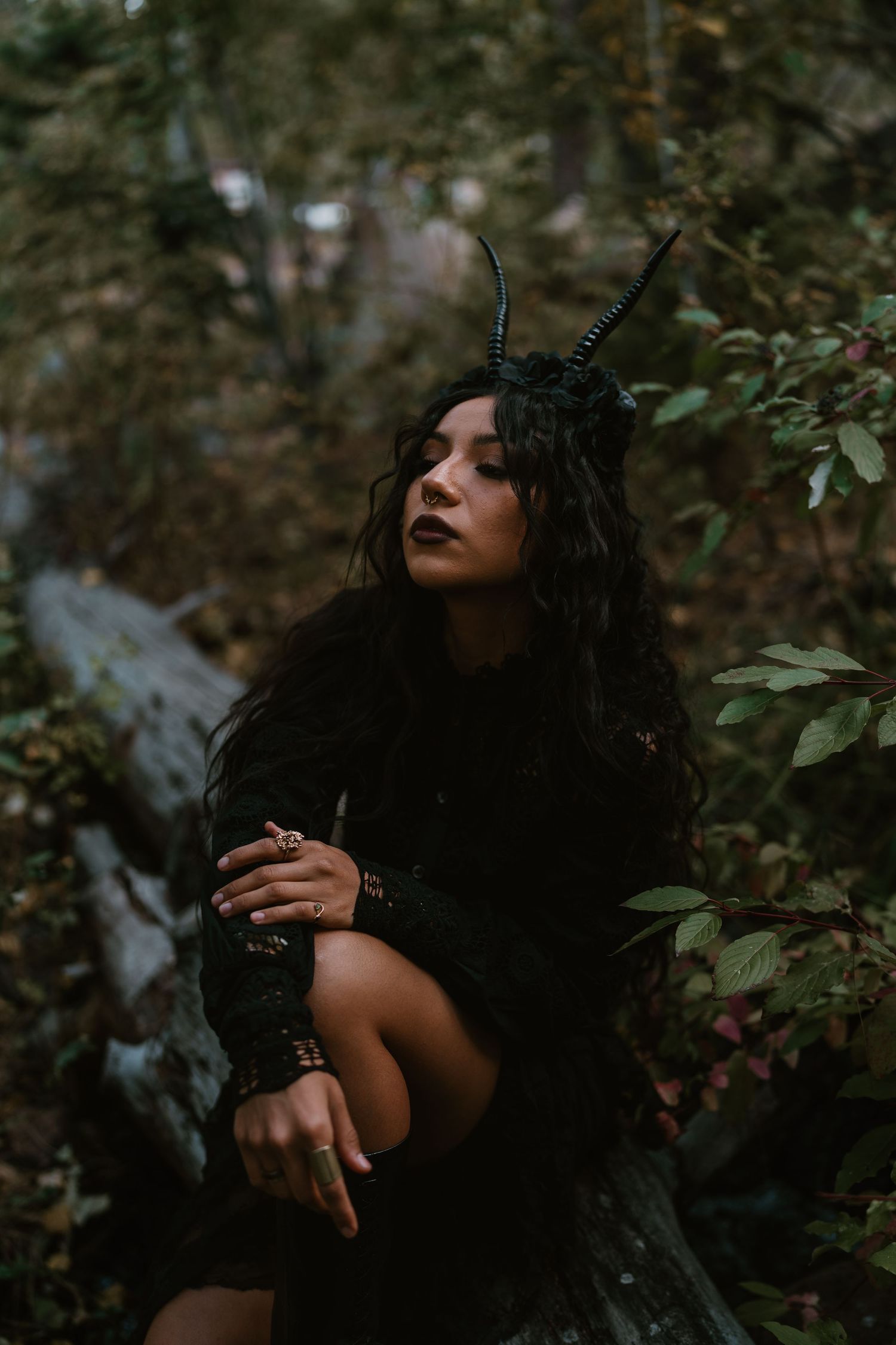 Woman in dark fantasy costume with black horns headpiece and gothic makeup poses mysteriously in a forest setting.