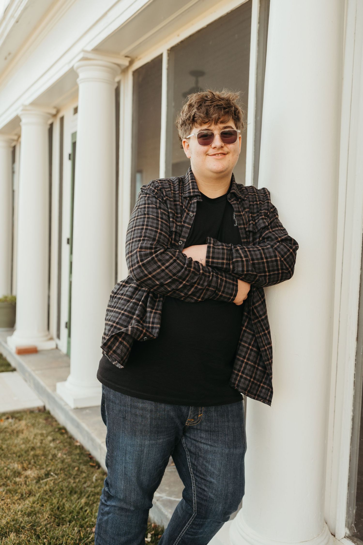 Person in sunglasses and plaid blazer poses casually against white columns in outdoor setting.