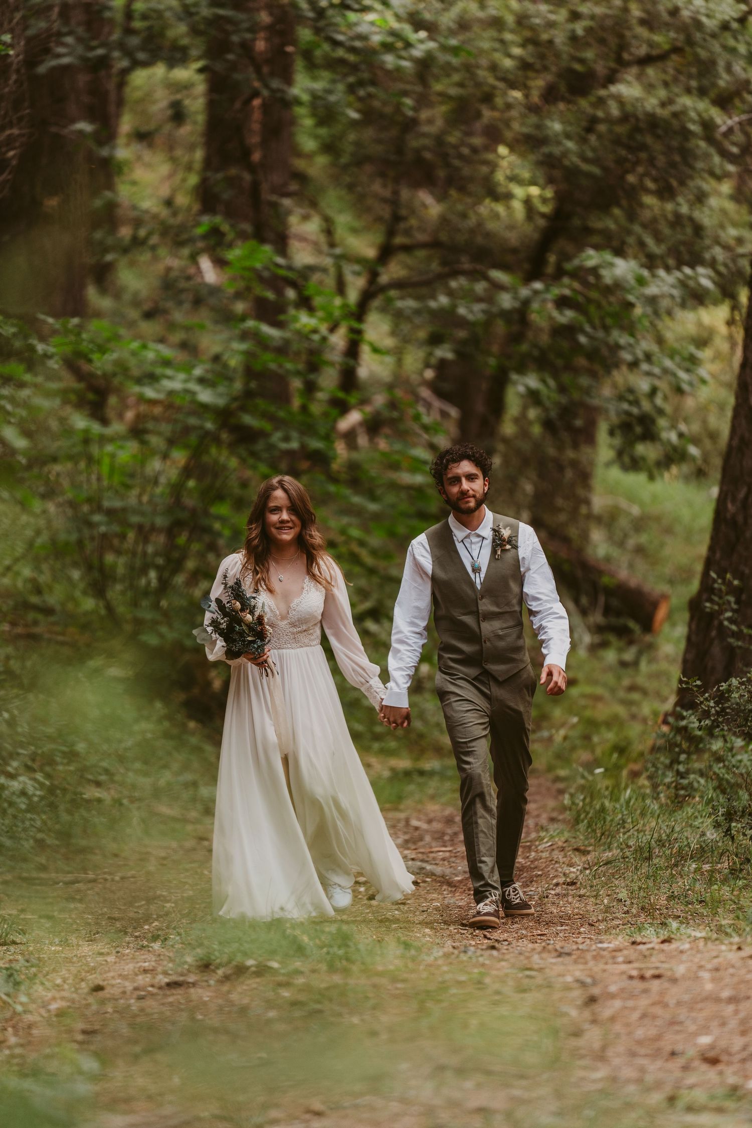 A couple walks hand in hand along a wooded forest path during their romantic outdoor wedding ceremony.