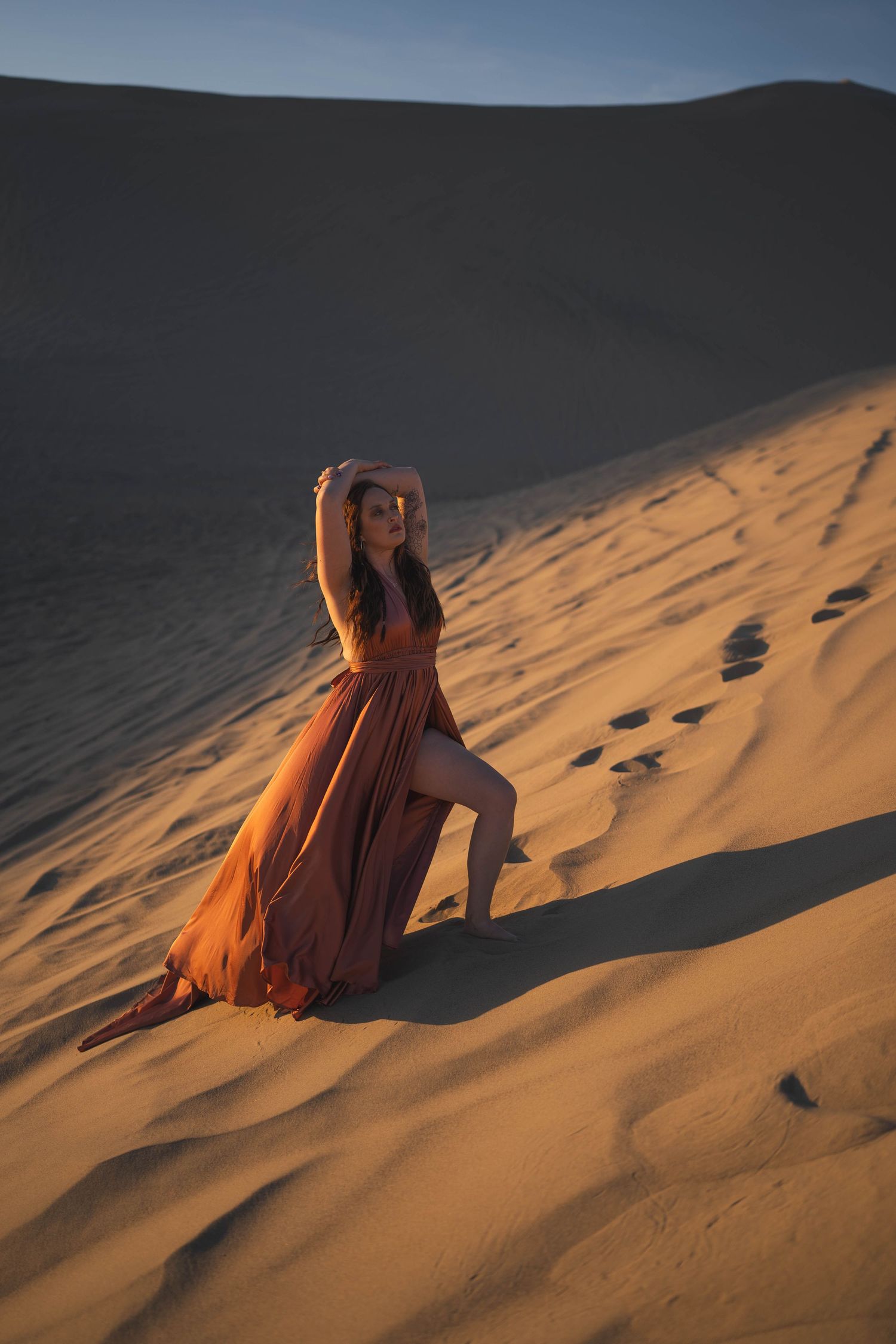 Figure in flowing orange dress poses dramatically on sand dunes at sunset casting long shadows in desert landscape.