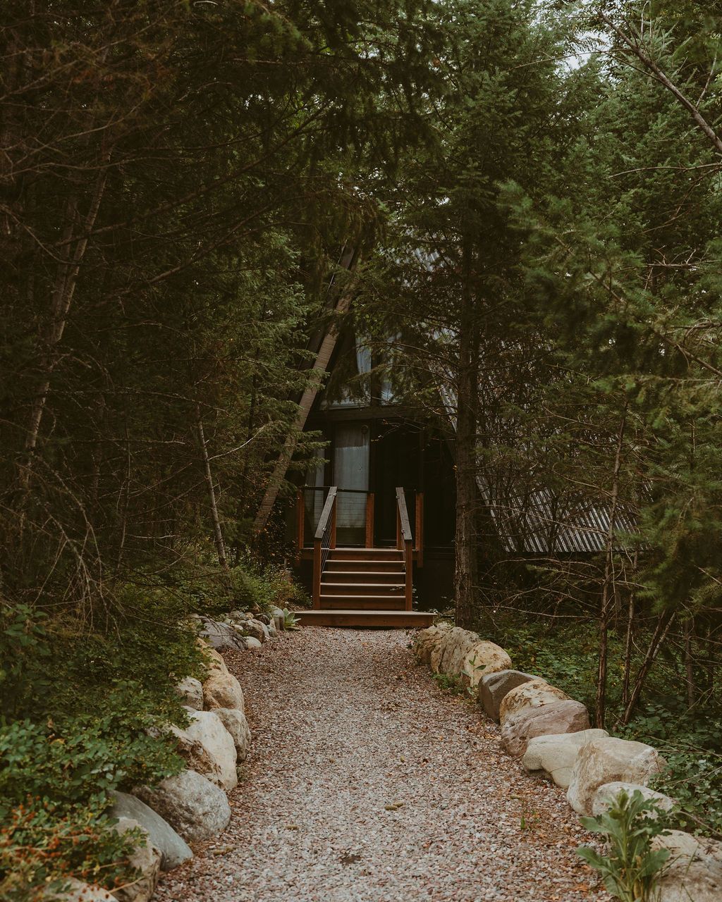 A-frame cabin in the woods with a gravel path lined by large rocks leading to wooden entry stairs.
