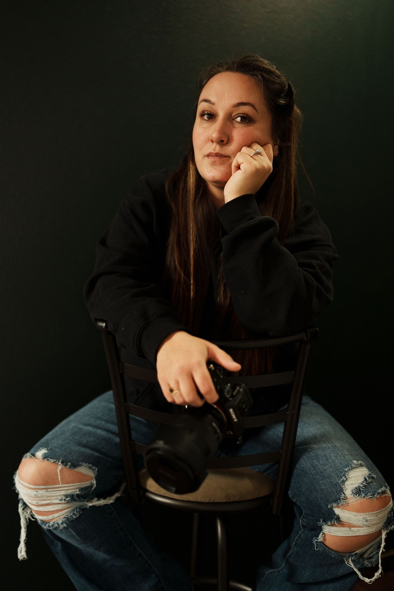 Person in ripped jeans and black sweater poses against dark background in artistic portrait series.