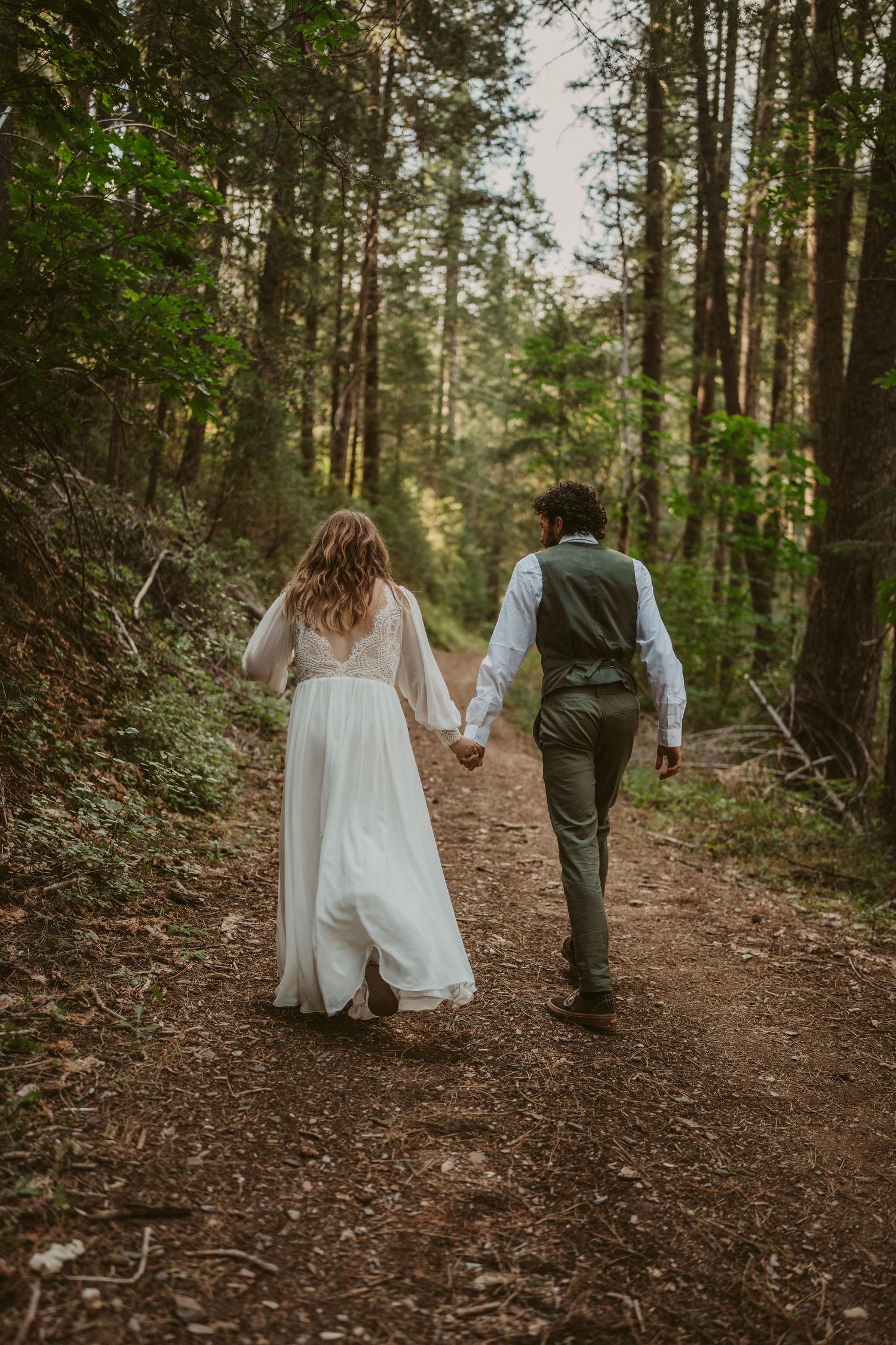 A couple in wedding attire walks hand in hand down a forest trail surrounded by tall evergreen trees.