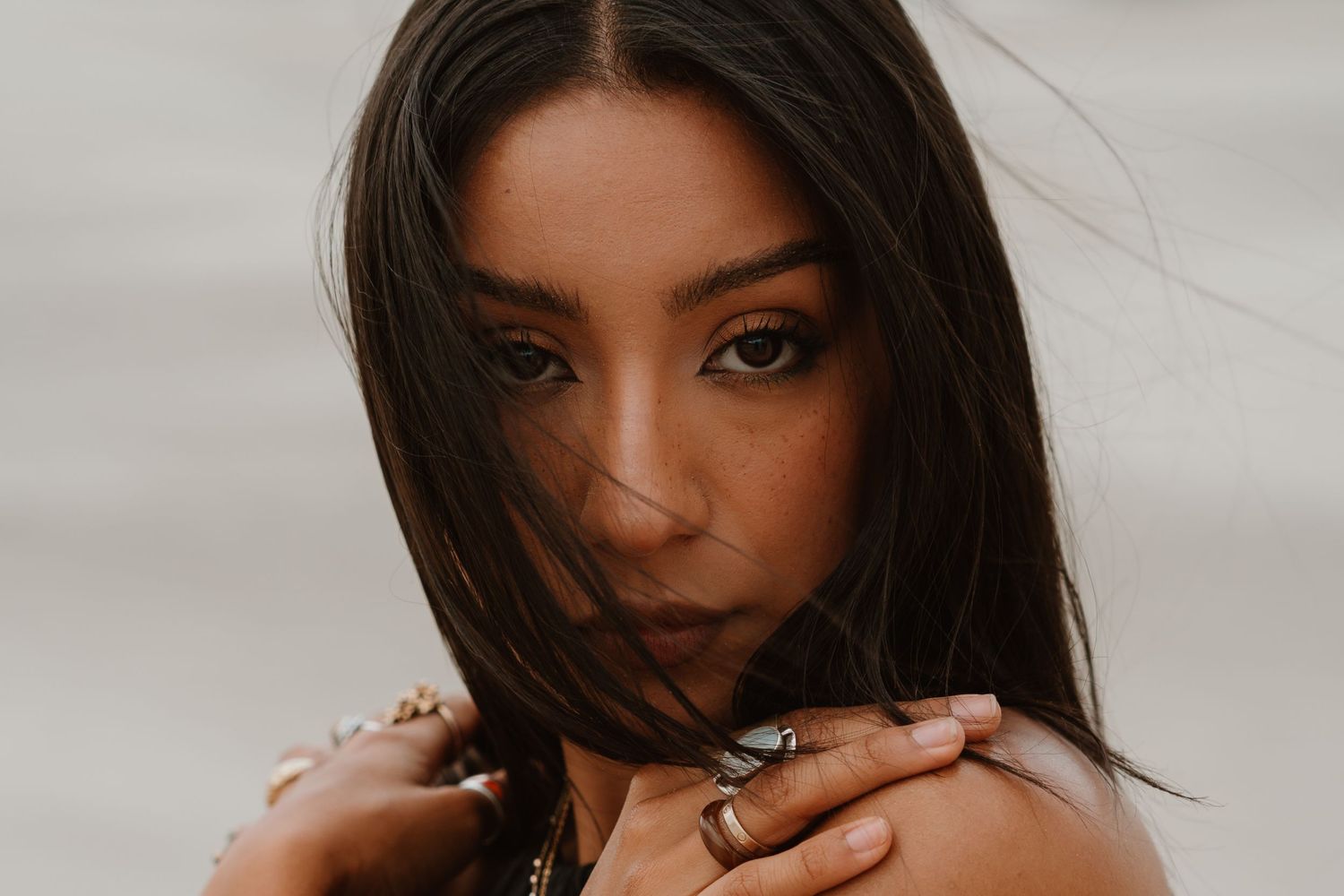 Young woman with dark hair and rings posing outdoors, wind blowing hair across her face.
