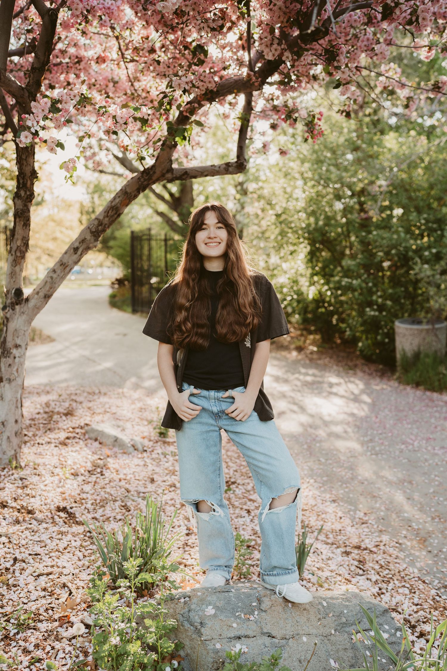 Young woman in black tee and ripped jeans smiling under pink cherry blossom tree on a sunny spring day.