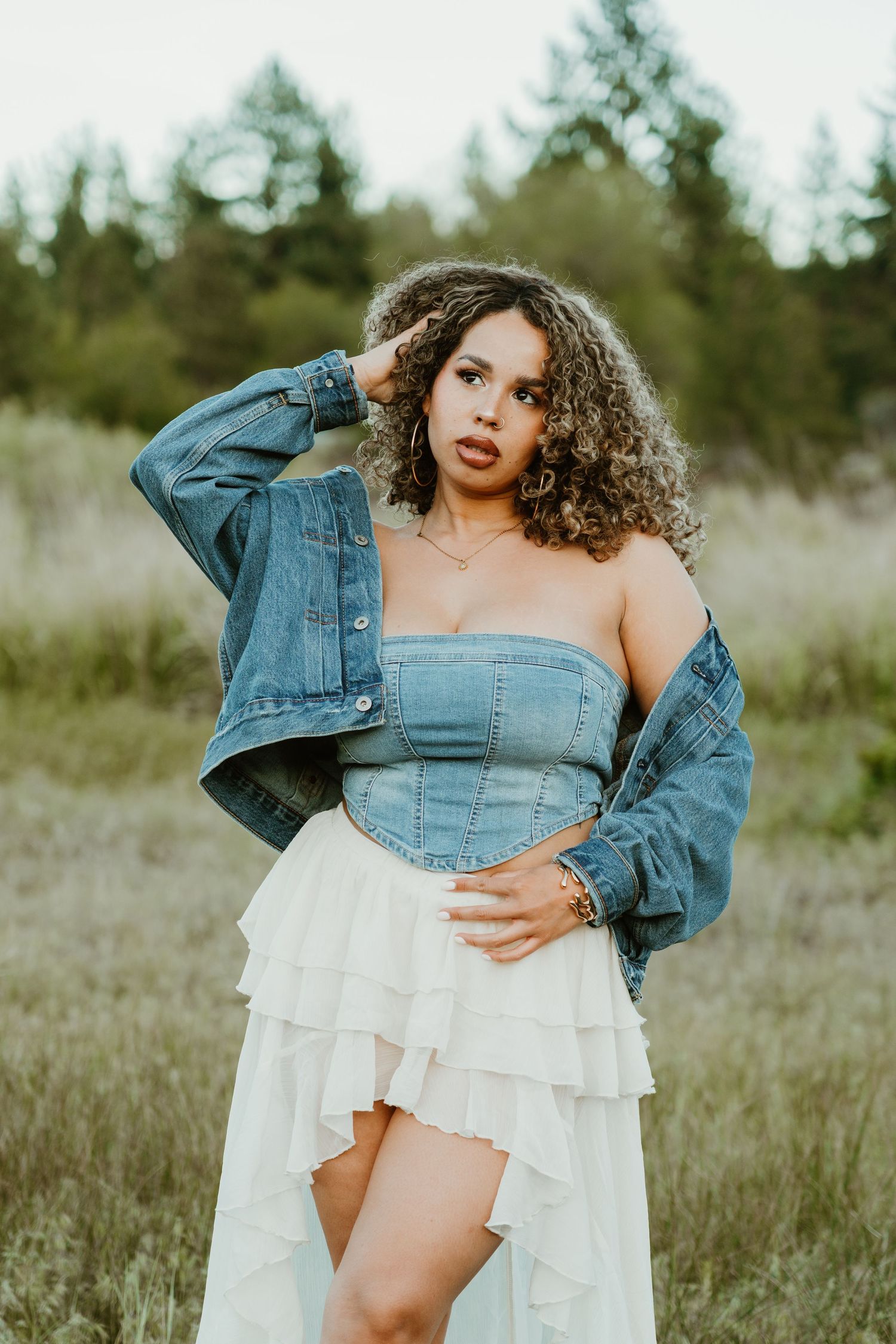 Woman in denim corset top and white ruffle skirt poses in a field wearing an oversized denim jacket.