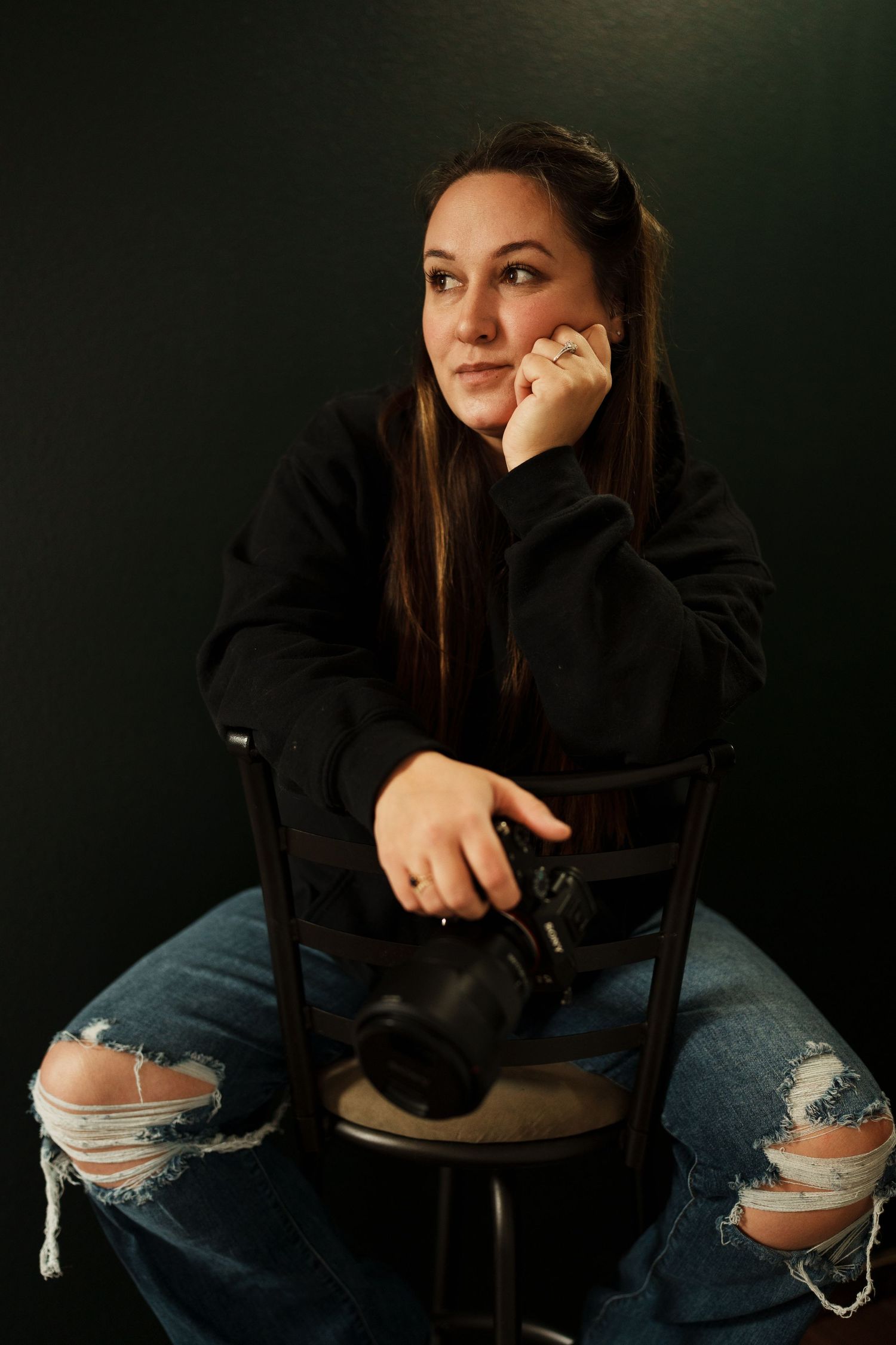Person in black hoodie and ripped jeans sits in moody lighting against dark background.