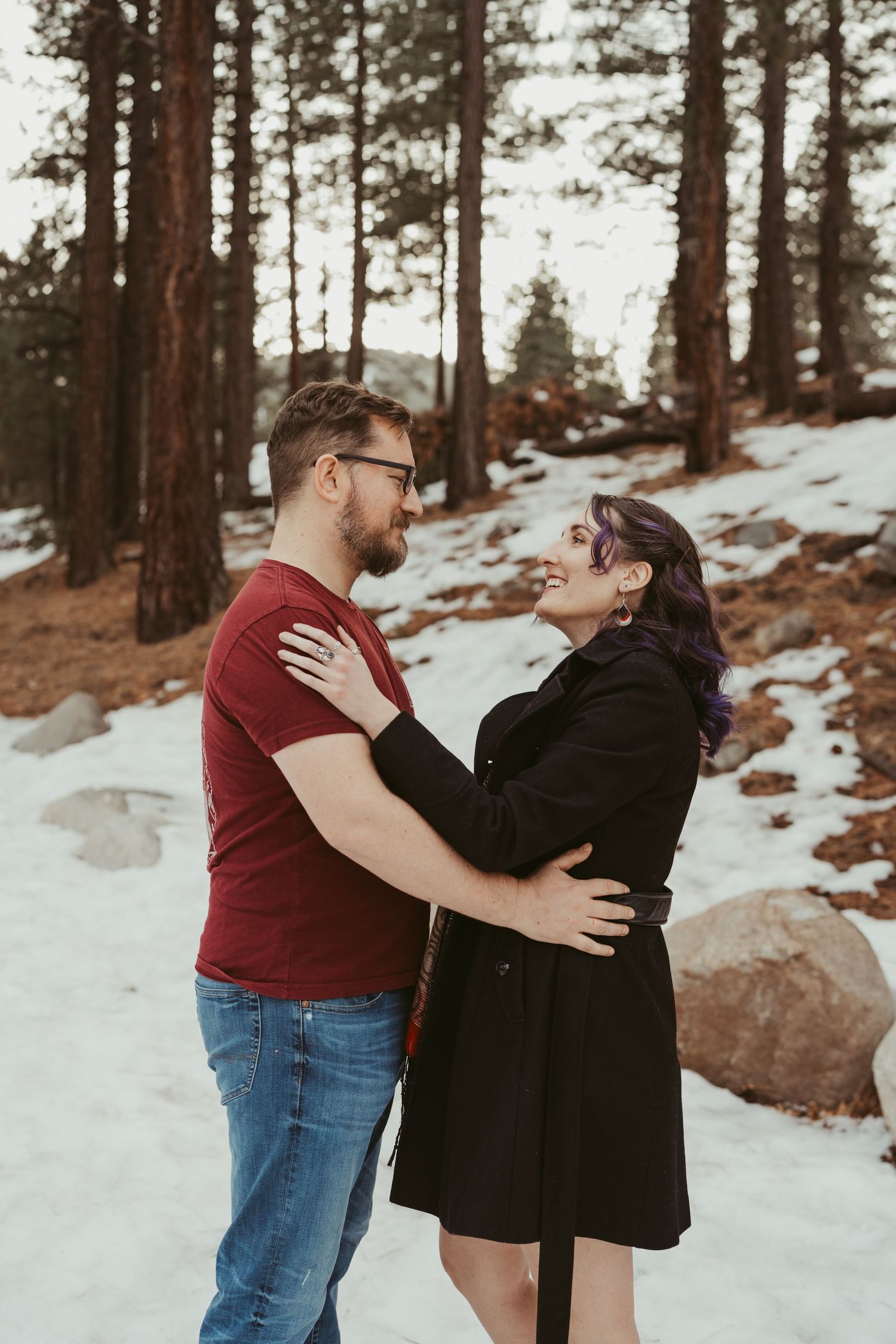 A couple embraces in a snowy forest setting with tall pine trees and rocky terrain in the background.