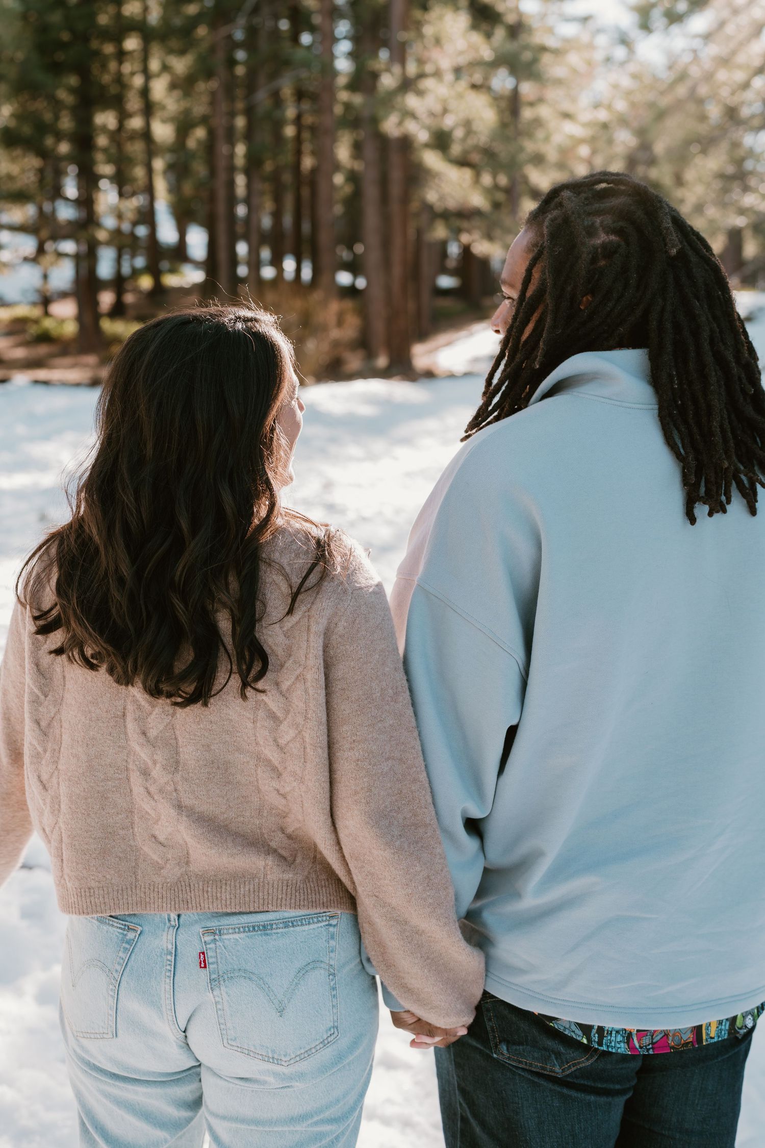 A couple in casual winter wear holding hands while walking through a snowy forest path.