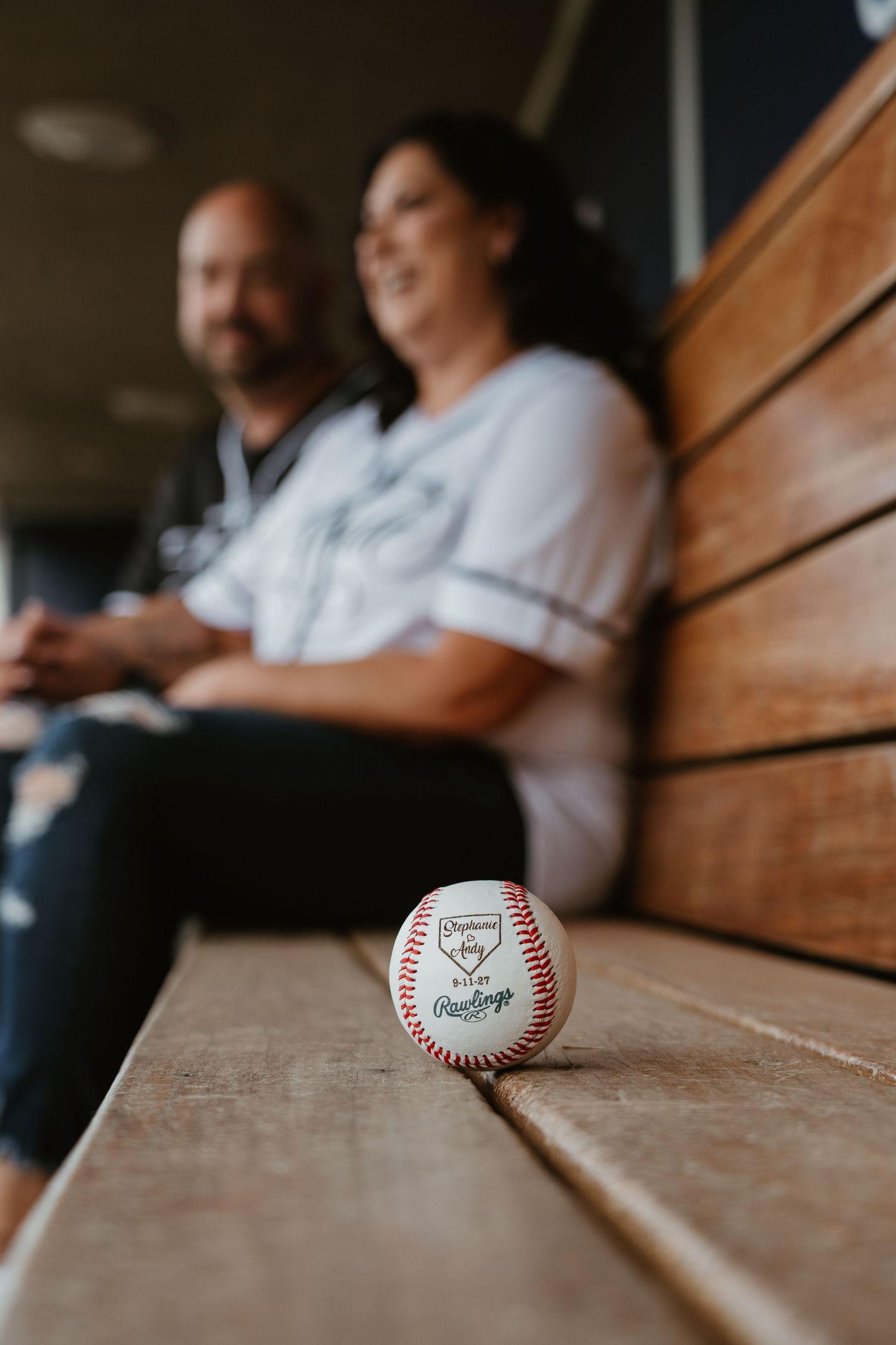 Rawlings baseball on dugout bench with couple in baseball jerseys sitting blurred in background.