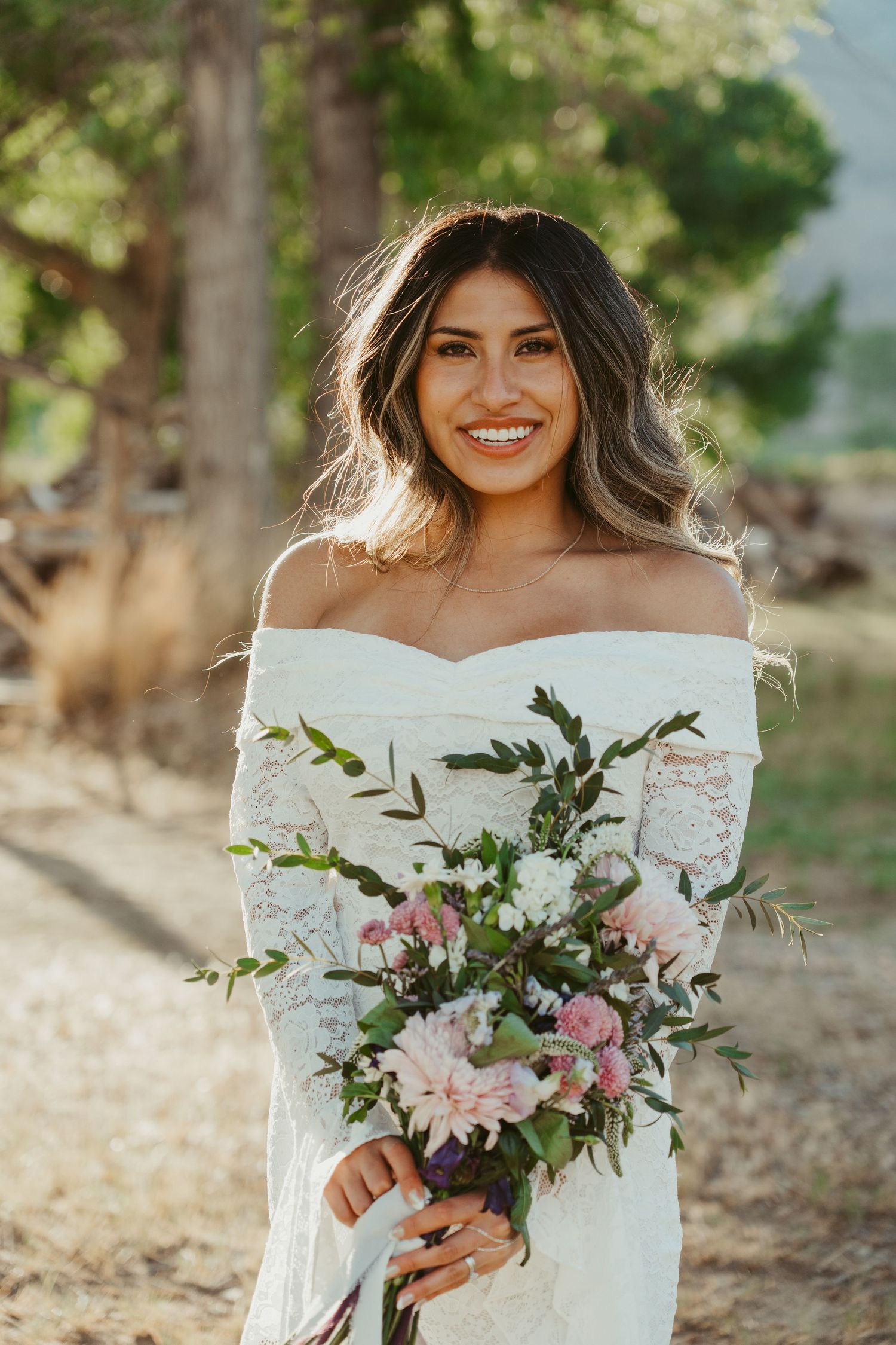 Smiling bride in off-shoulder lace gown holding pink floral bouquet outdoors among trees.