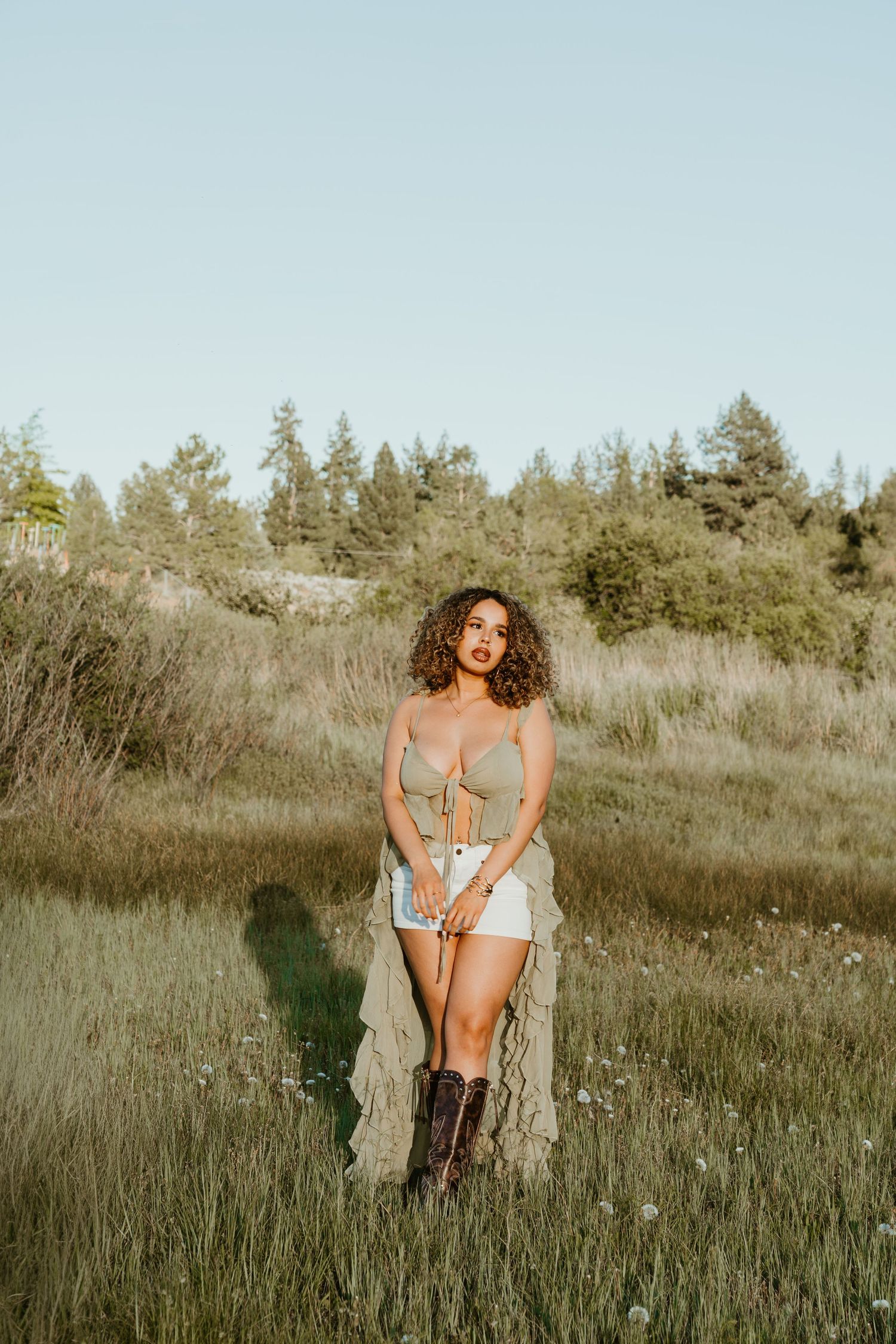 Woman in crop top, denim skirt, and cowboy boots posing in a scenic meadow with trees in the background.