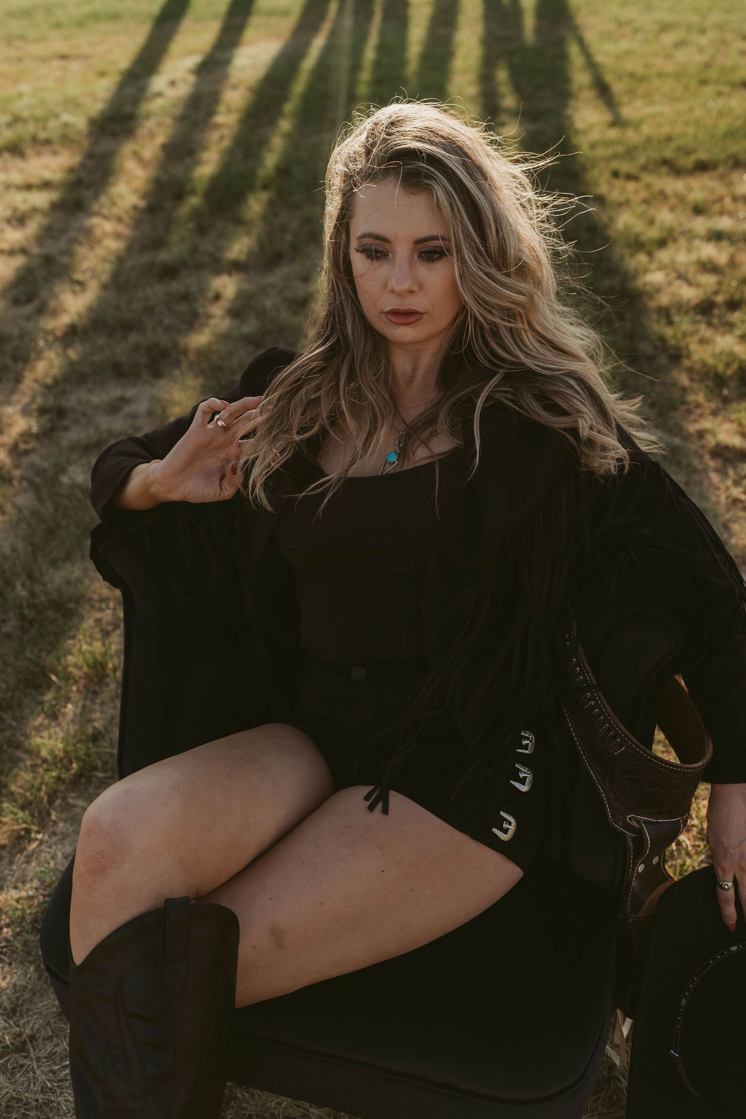 Artistic fashion photo of someone in a black outfit posed outdoors in arm chair during golden hour with long shadows on grass.