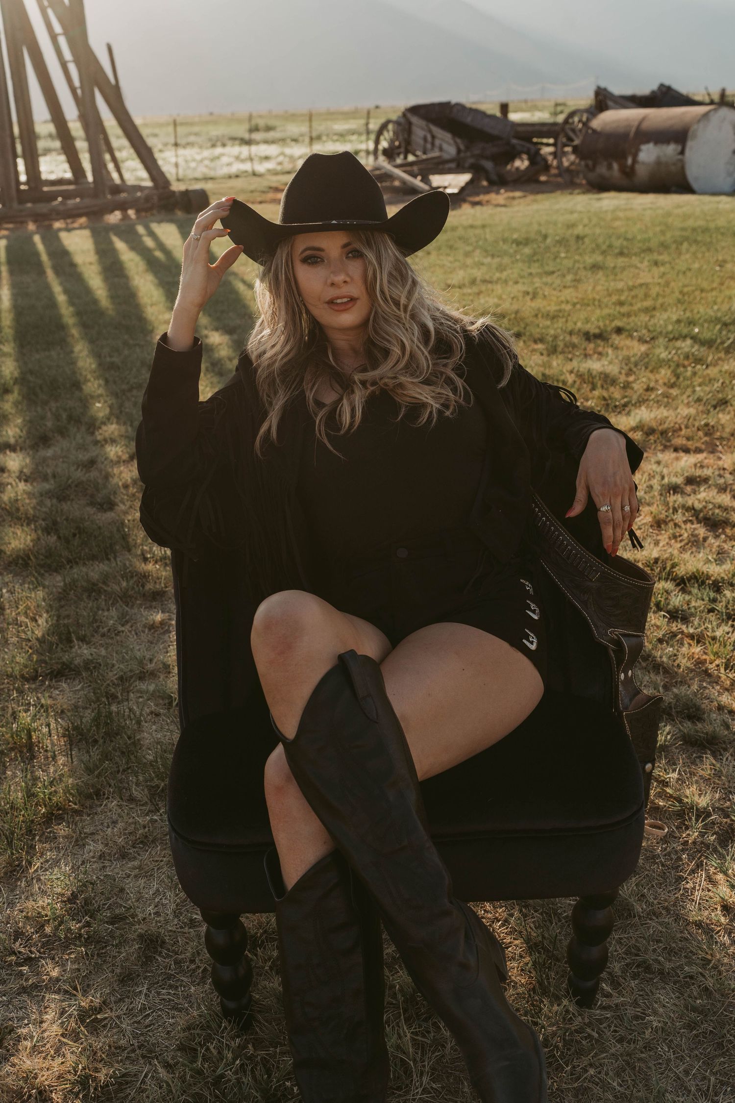 Style photo of western fashion with black boots, cowboy hat and fringed black jacket captured at golden hour in a rustic field setting.