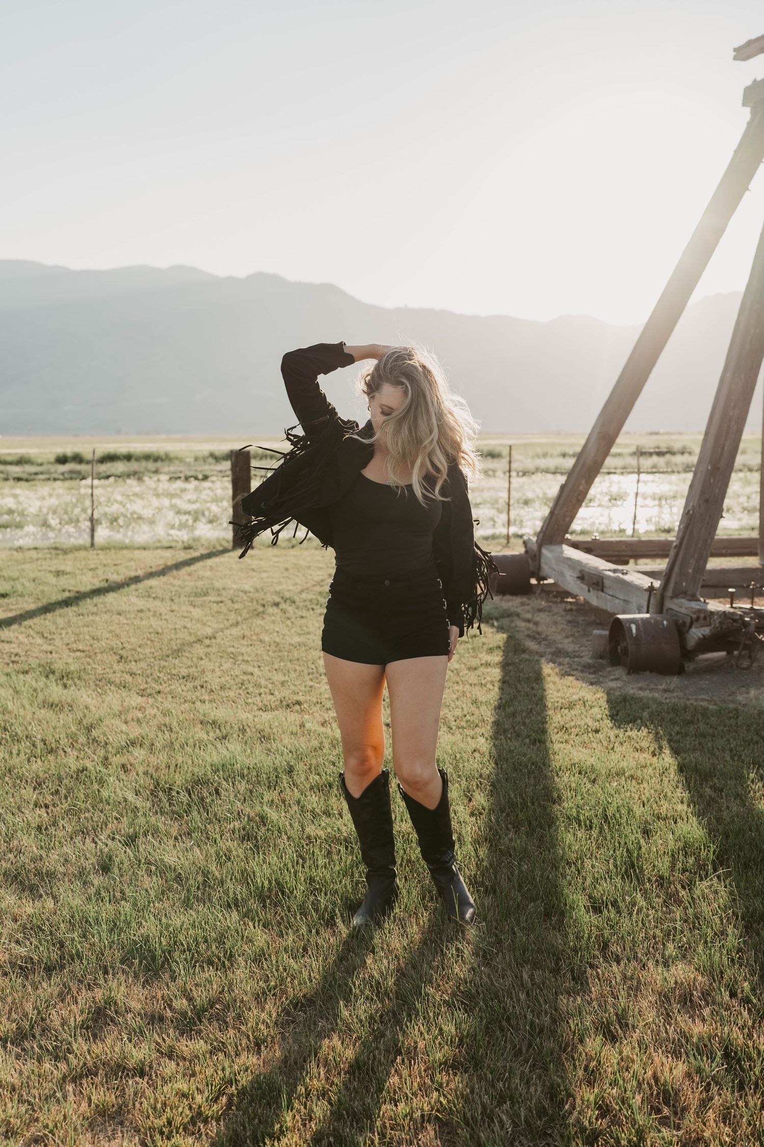 Silhouette against mountain landscape at sunset with black boots and modern western wear casting long shadows on grassy field.
