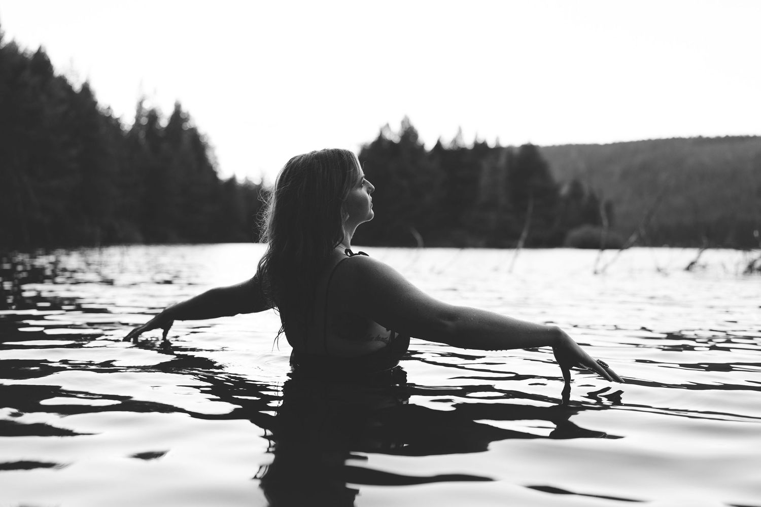 Black and white photo of a woman gracefully posing in a calm lake surrounded by forest trees.