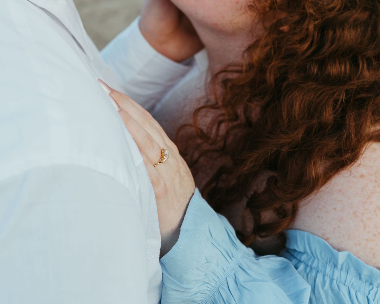 A close-up view of someone wearing a light blue shirt with curly red hair holding on to her newly engaged fiance.