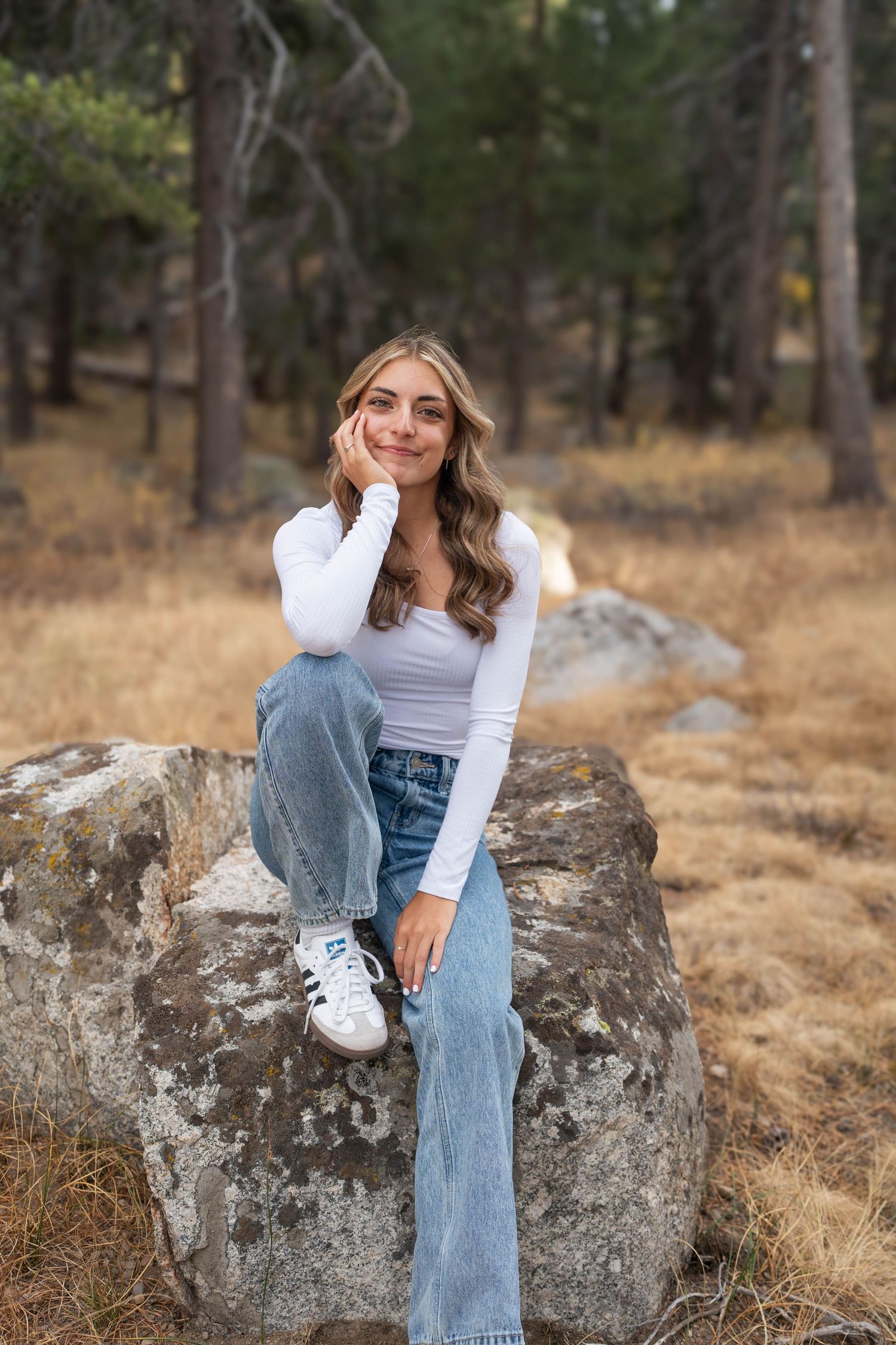 Person in light colored top and jeans sits casually on large boulder in outdoor woodland setting during autumn.