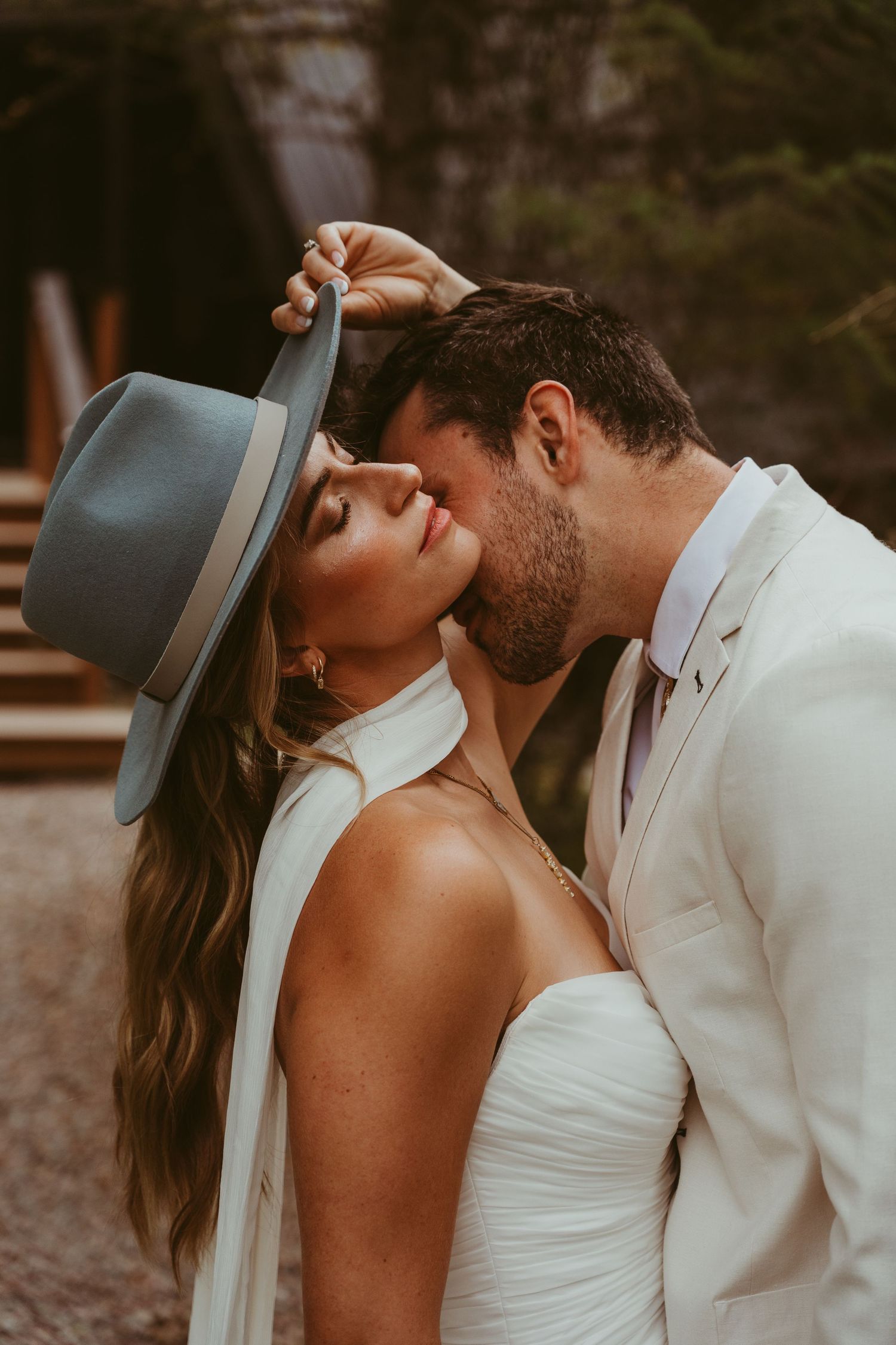 A romantic wedding couple shares an intimate moment outdoors as the bride in white holds a gray felt hat.