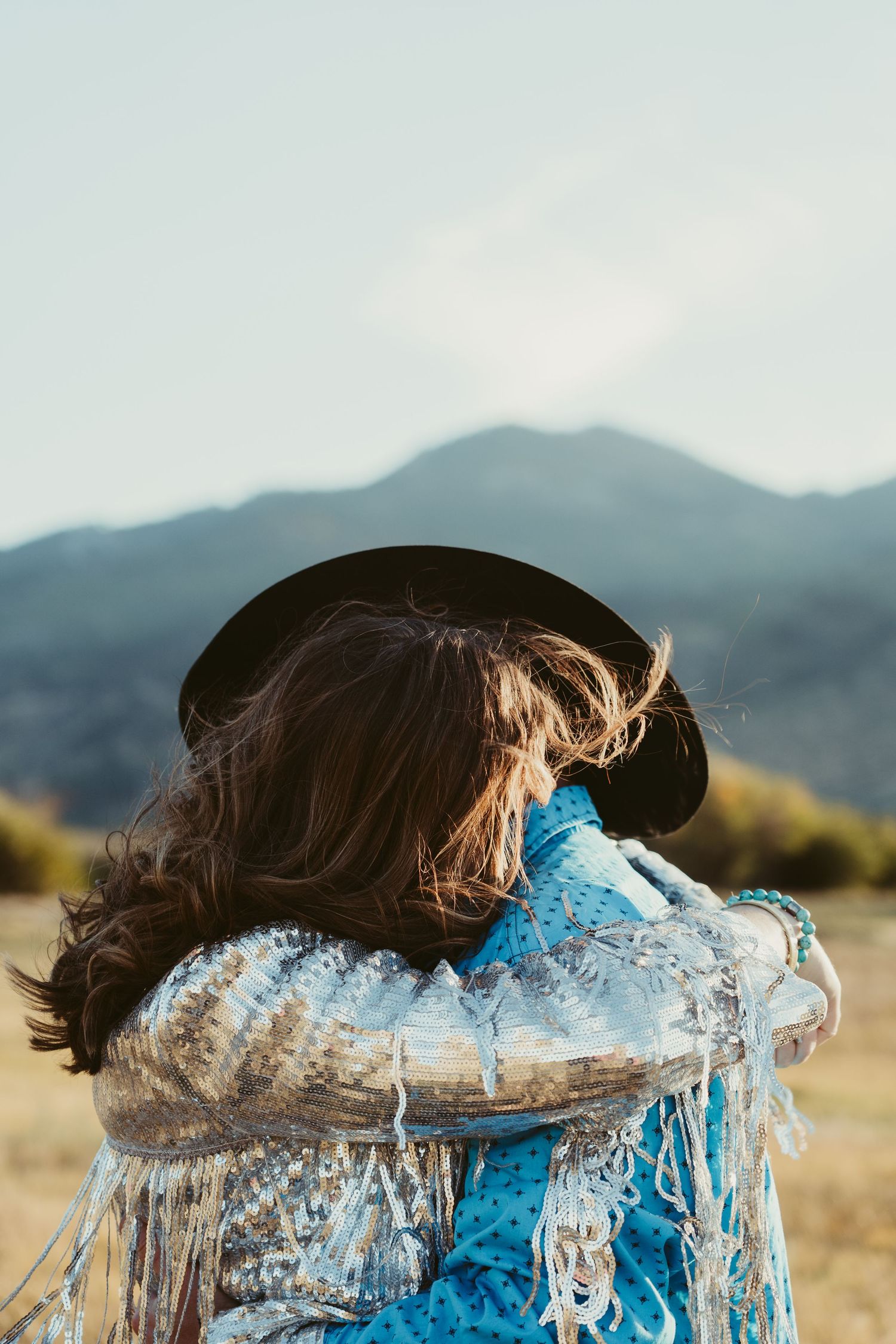 Woman in bohemian blue fringe jacket and black hat standing in a field with mountains in the background.