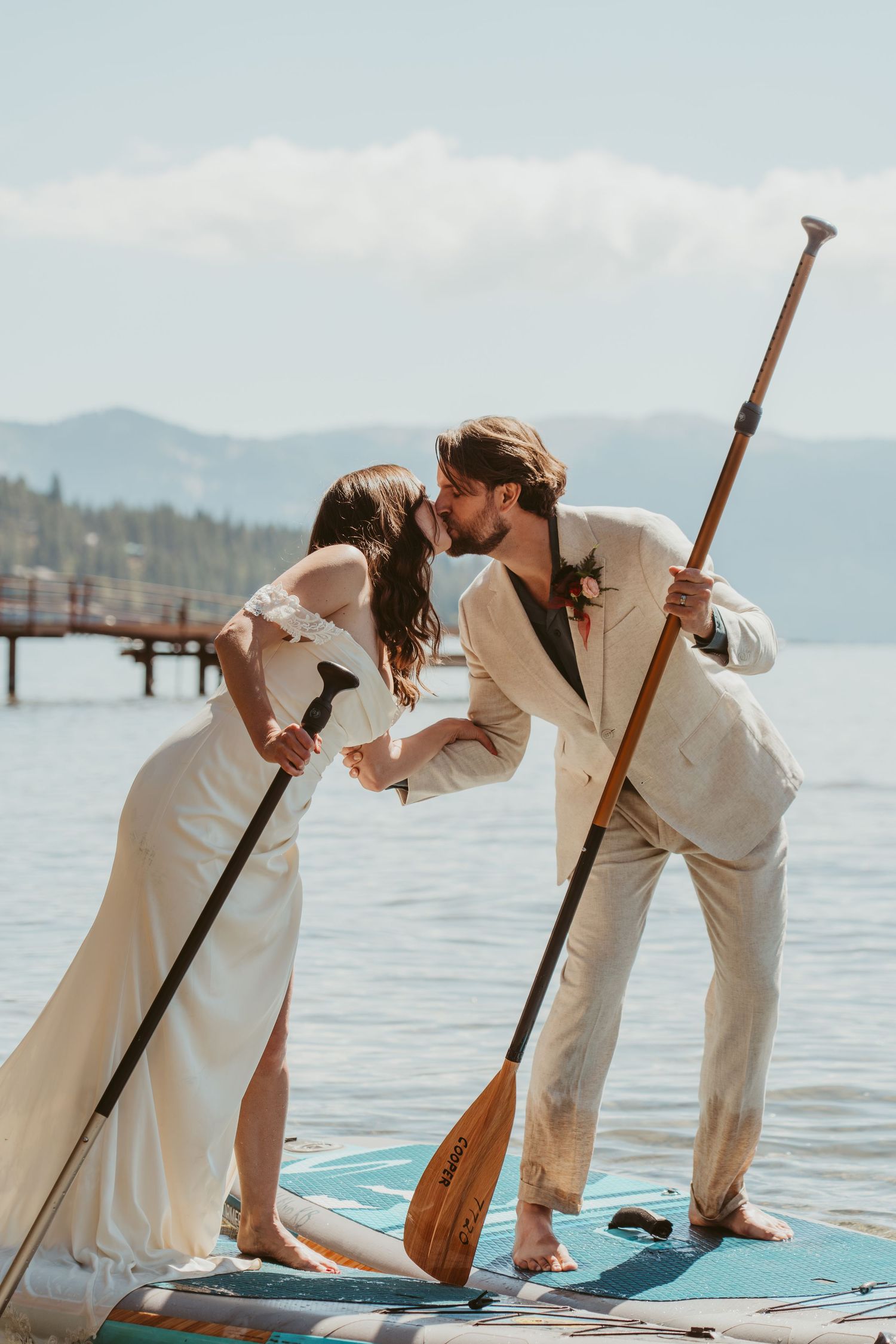 Couple shares romantic kiss while paddleboarding on lake with mountain backdrop and wooden pier in distance.