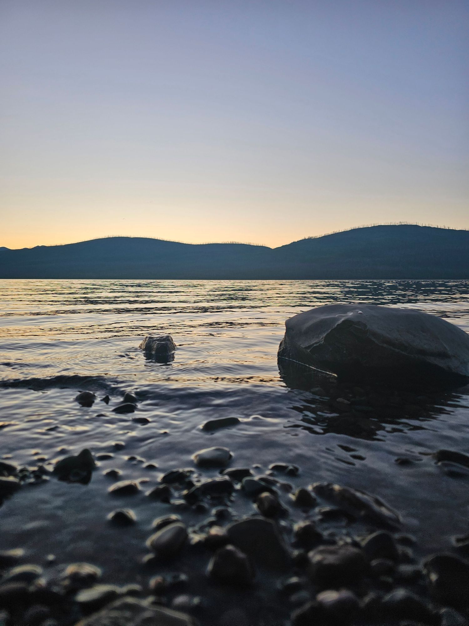 Sunset silhouettes rocky shoreline with waves lapping against dark boulders as mountains fade into twilight horizon.