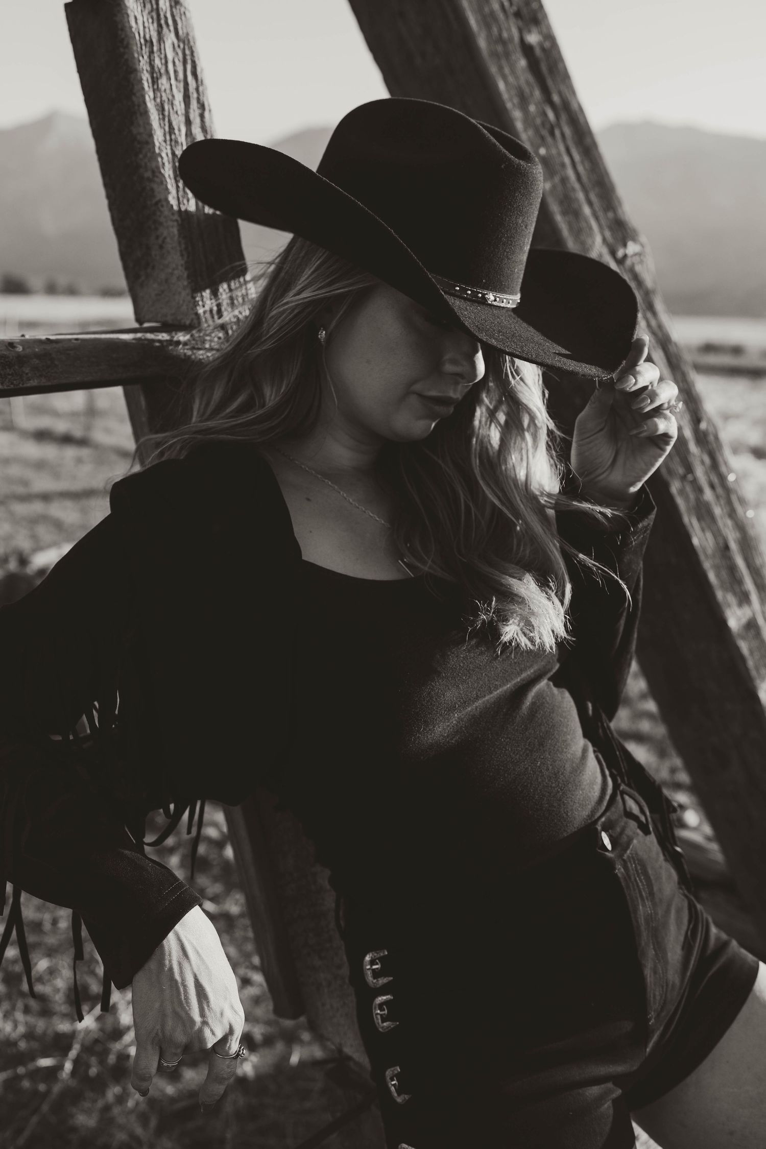 Dramatic black and white portrait of western cowgirl in black dress and wide-brimmed hat leaning against wooden fence.