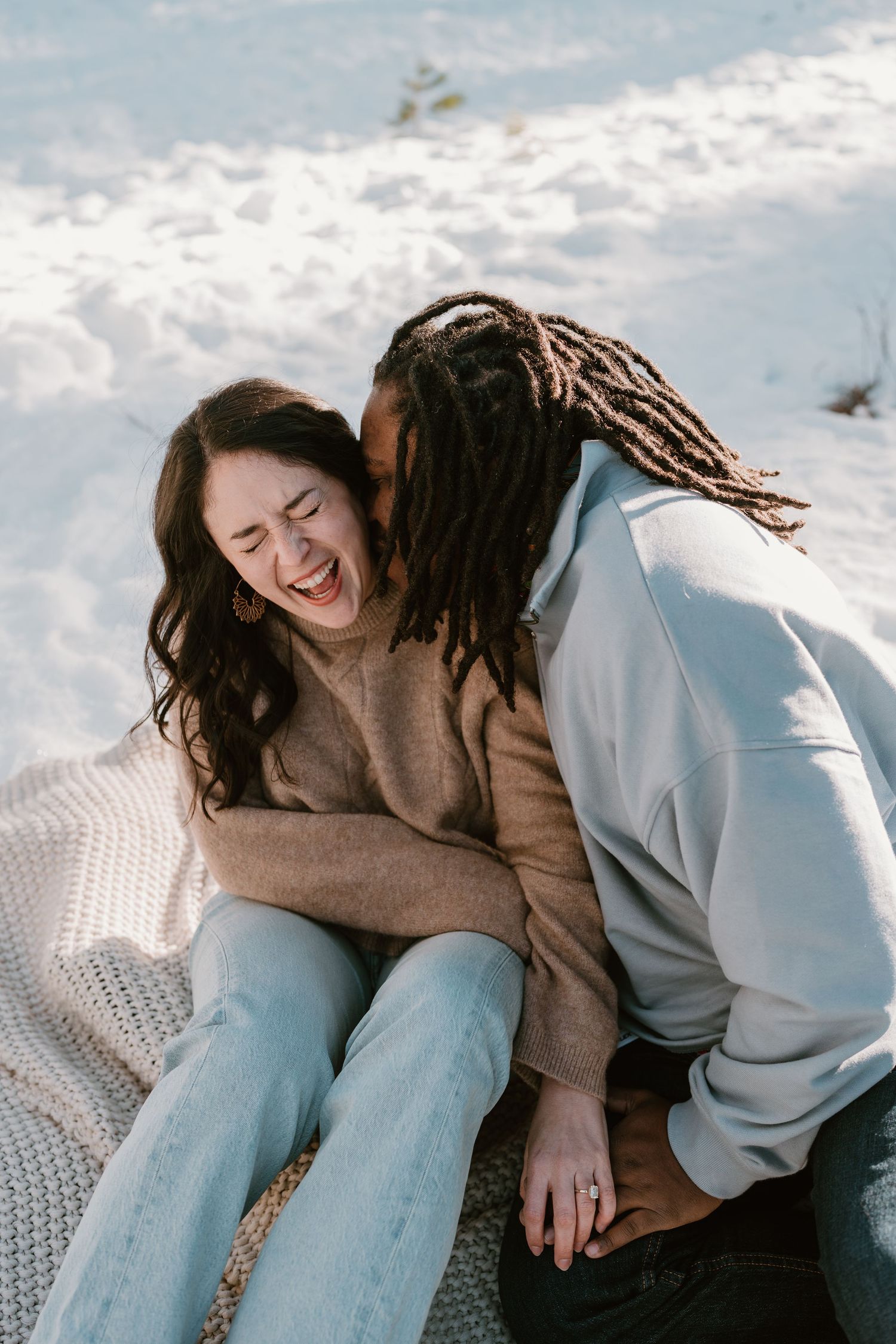 A joyful interracial couple laughing and sharing a tender moment together while sitting on a blanket in the snow.