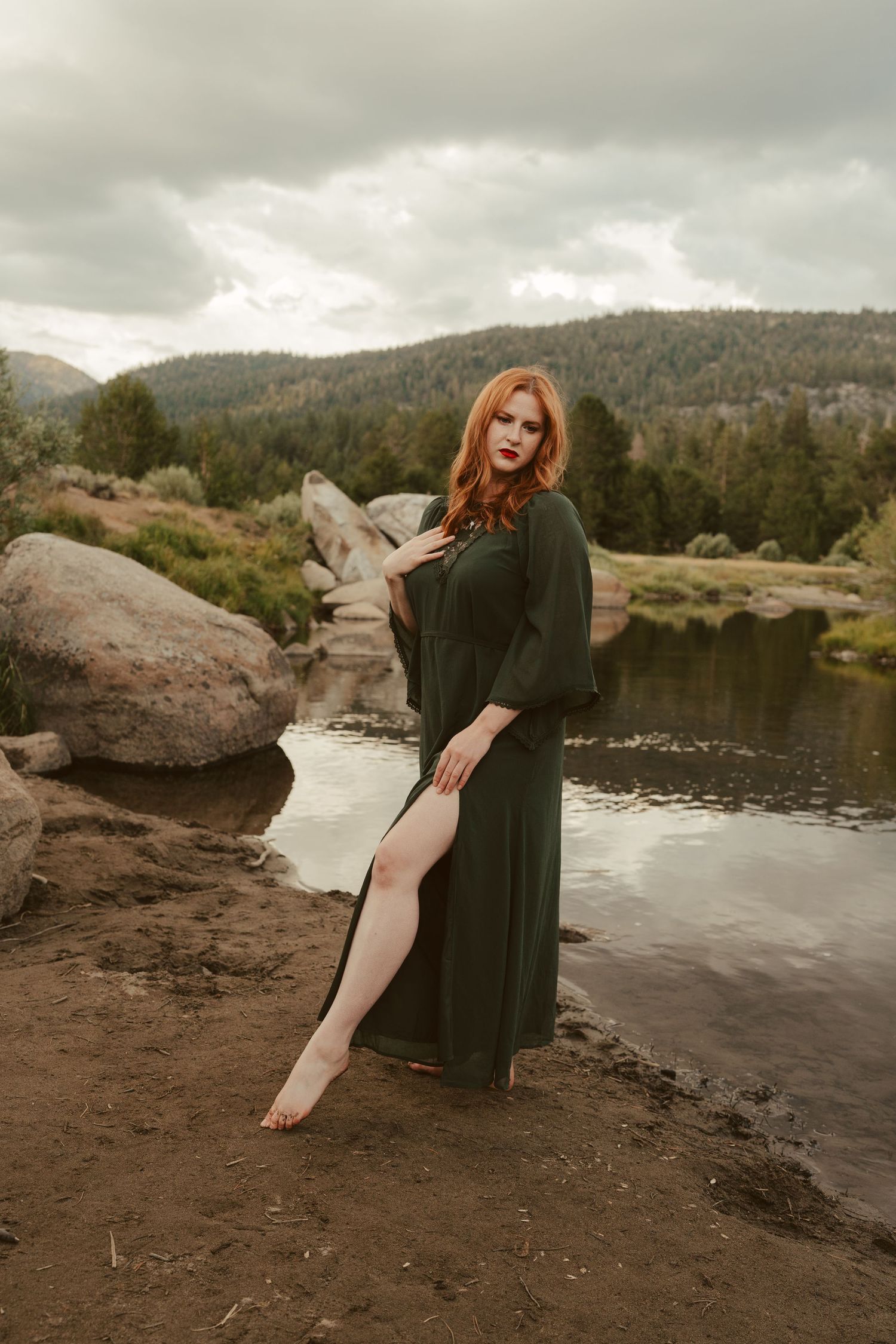 A person in a dark dress poses gracefully by a serene mountain lake surrounded by rocks and forest.