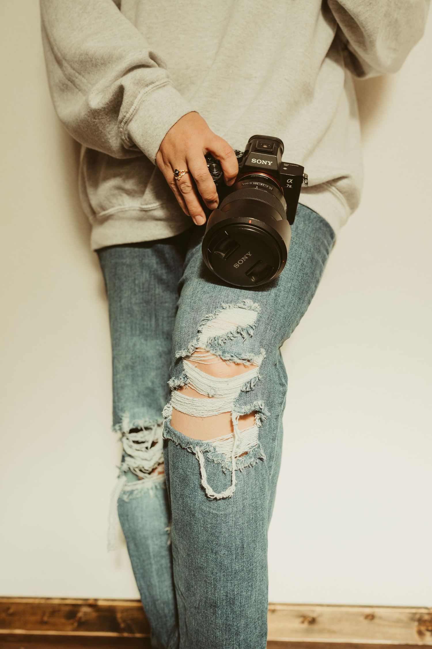 Person in ripped jeans and gray sweatshirt holding a Sony mirrorless camera against a white wall.